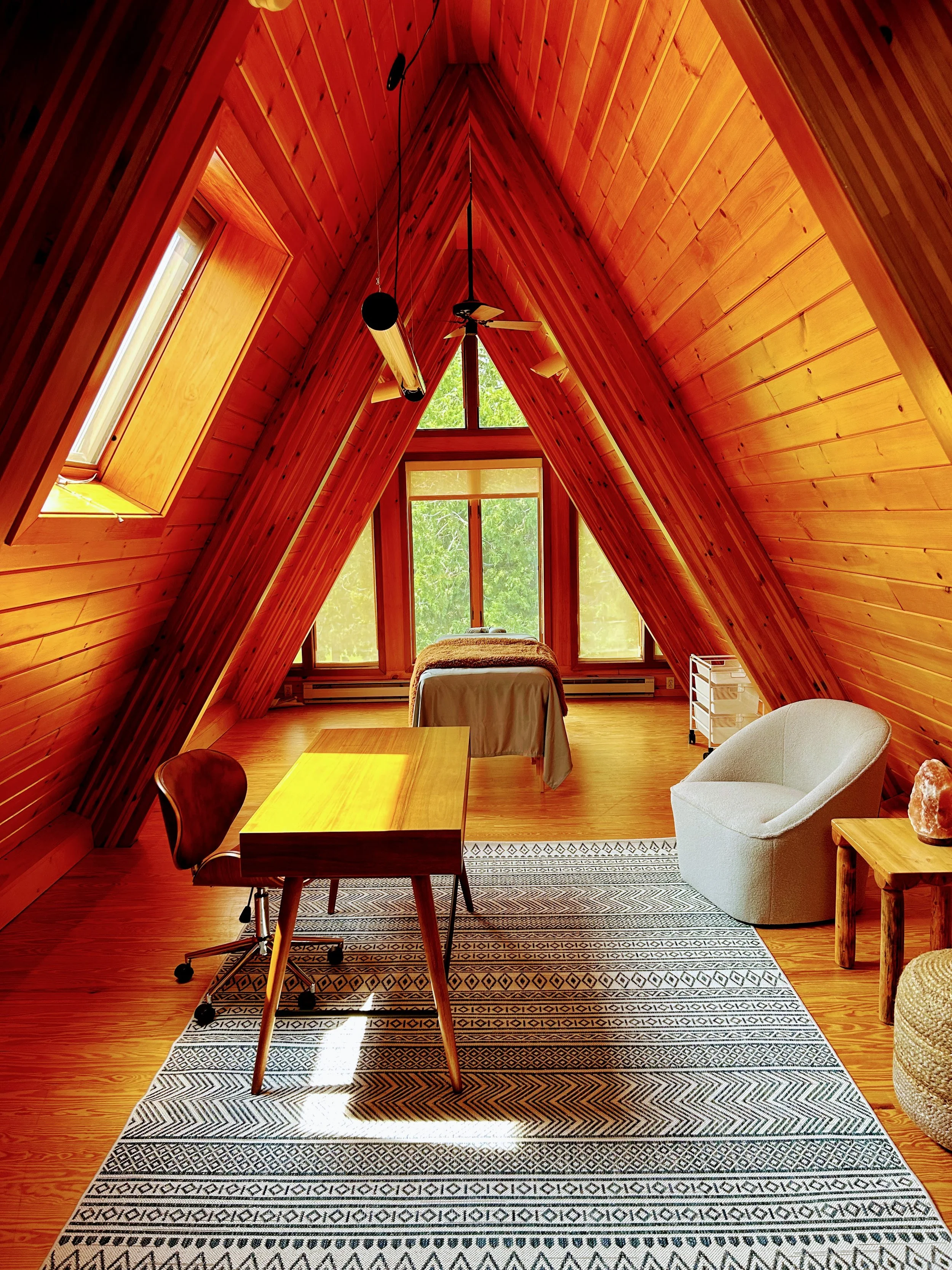 A-frame loft interior with wooden walls and ceiling, featuring a desk and chair on a patterned rug, a bed by the window, and a modern chair next to a side table.