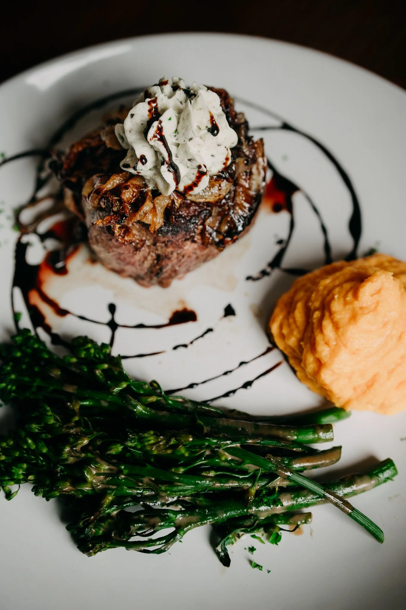 Gourmet steak dish with bleu cheese, mashed sweet potatoes, broccolini, and drizzle of balsamic glaze on a white plate.