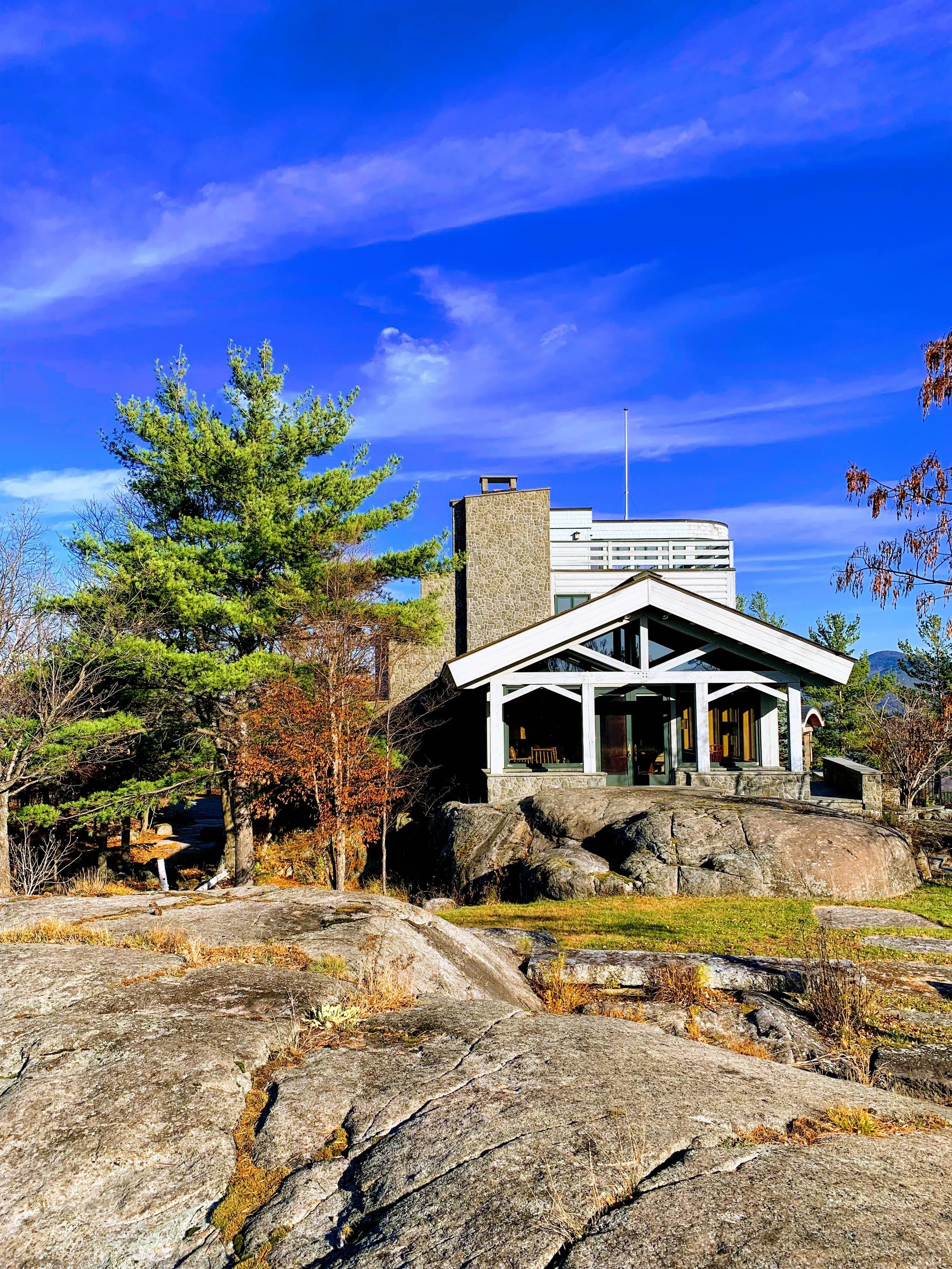 House with large windows on rocky hillside surrounded by trees under a blue sky.