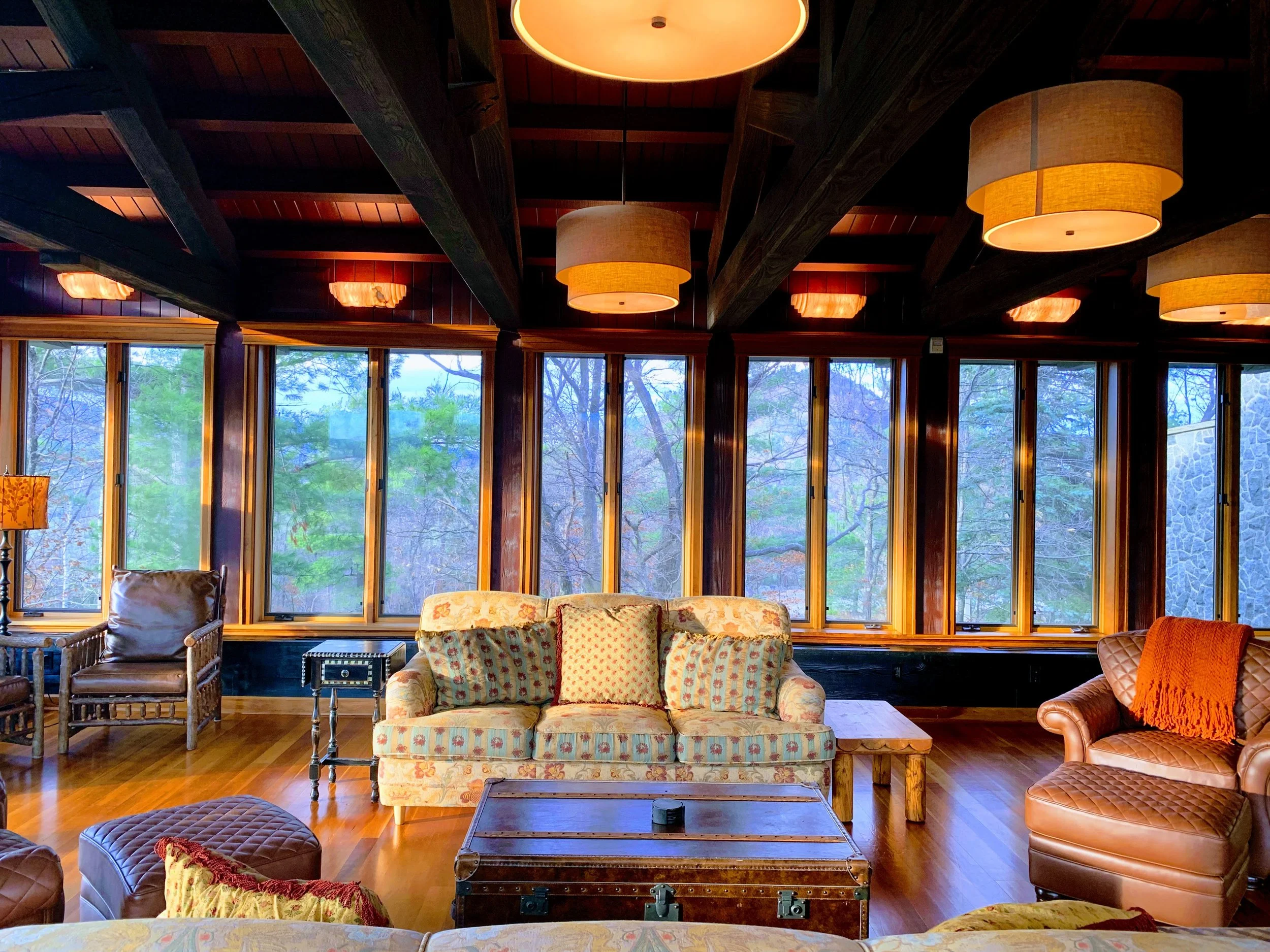 Cozy living room with large windows, floral sofa, armchairs, rustic wooden furniture, and warm lighting.
