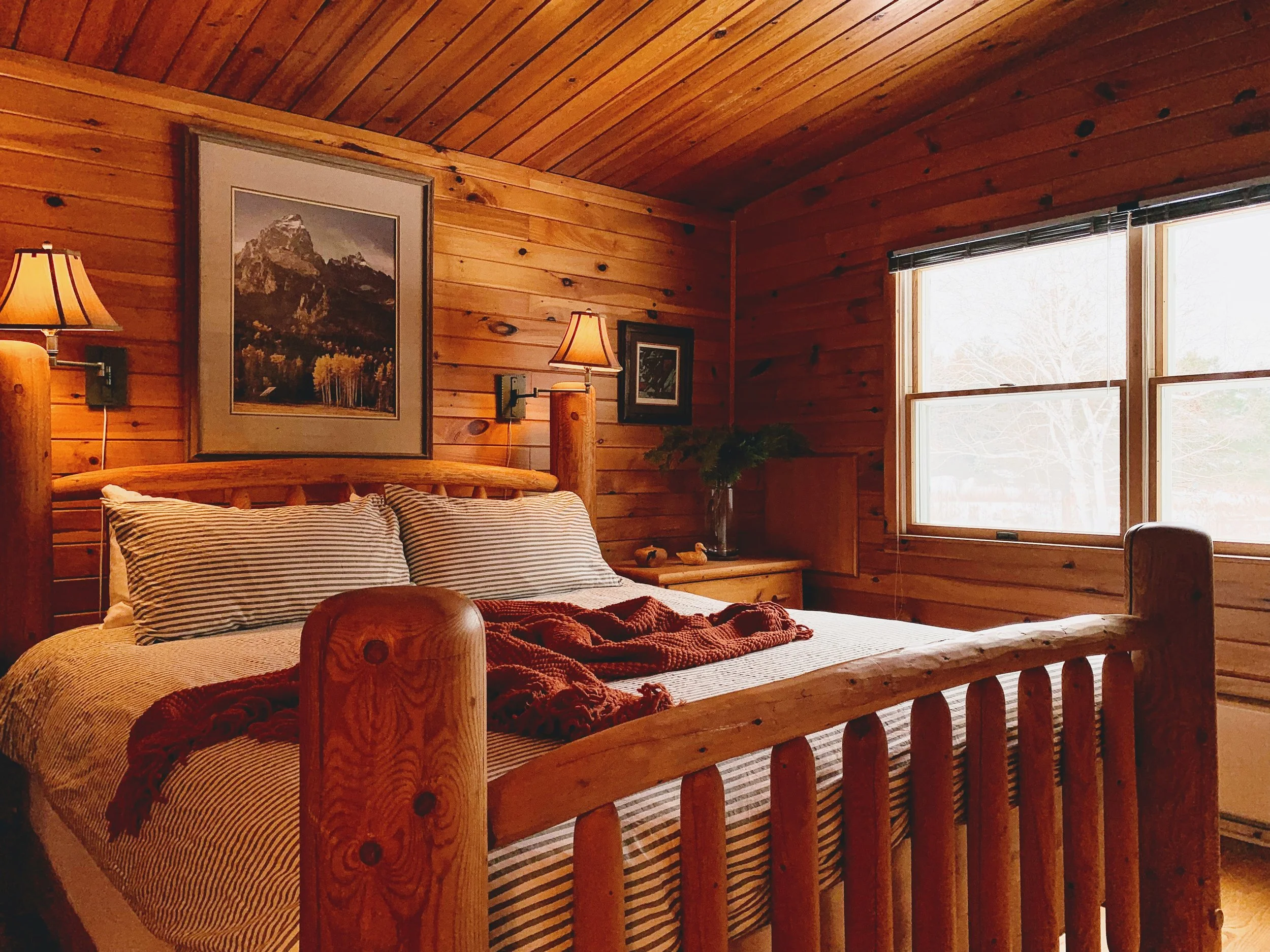Cozy rustic bedroom with wooden walls, a wooden bed frame, striped bedding, a red throw blanket, wall-mounted lamps, large picture window, and mountain landscape artwork.