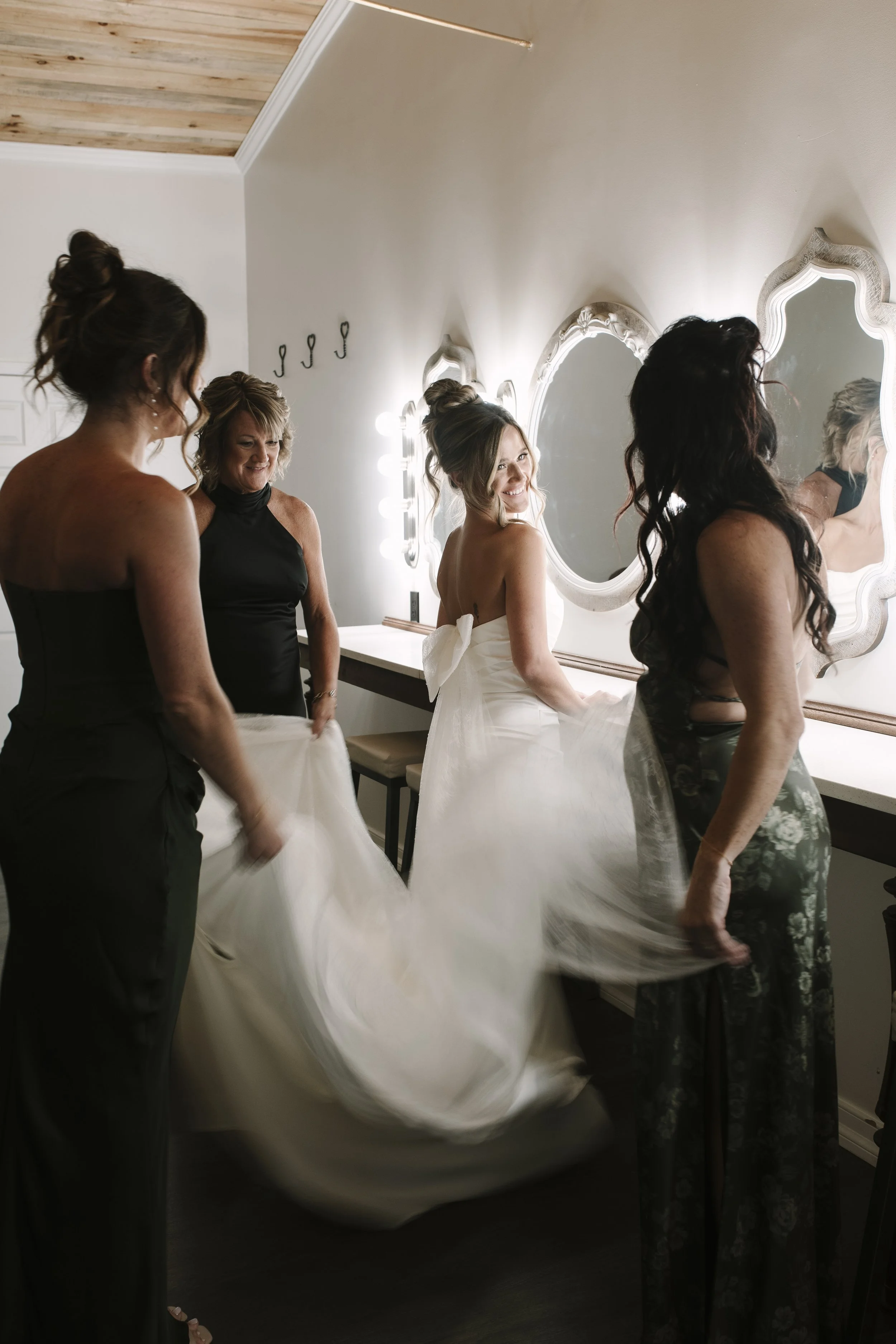 Bride in a white wedding dress surrounded by bridesmaids in a getting-ready room with ornate mirrors