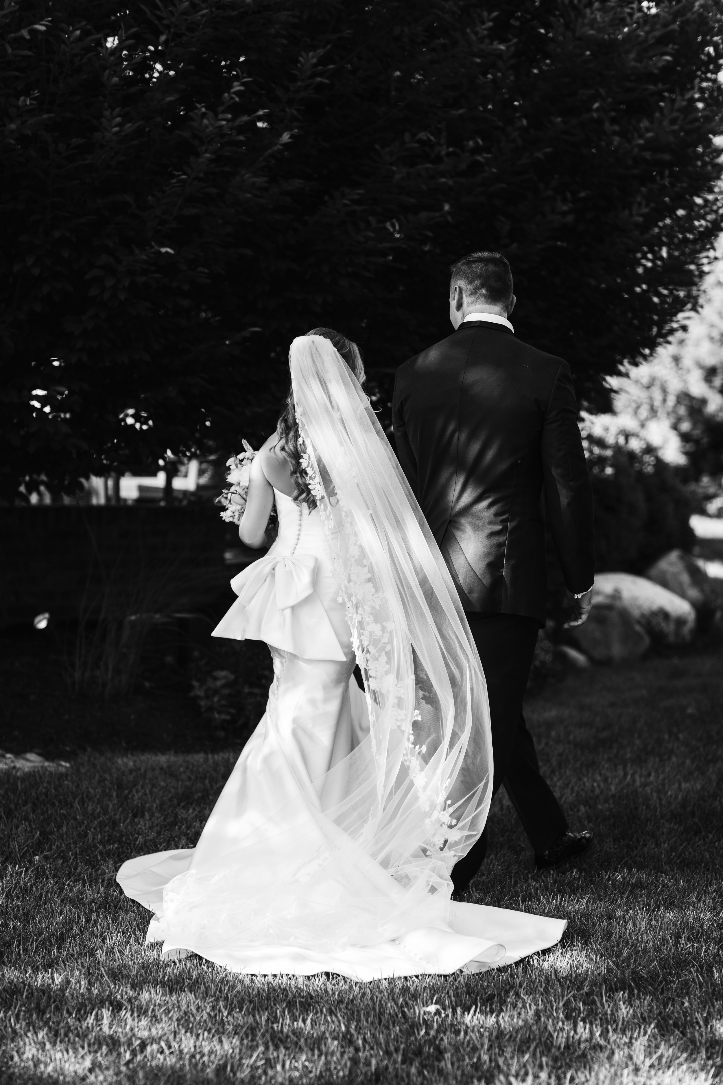 Black-and-white wedding portrait of a bride in a long veil and groom in a suit walking through a garden.
