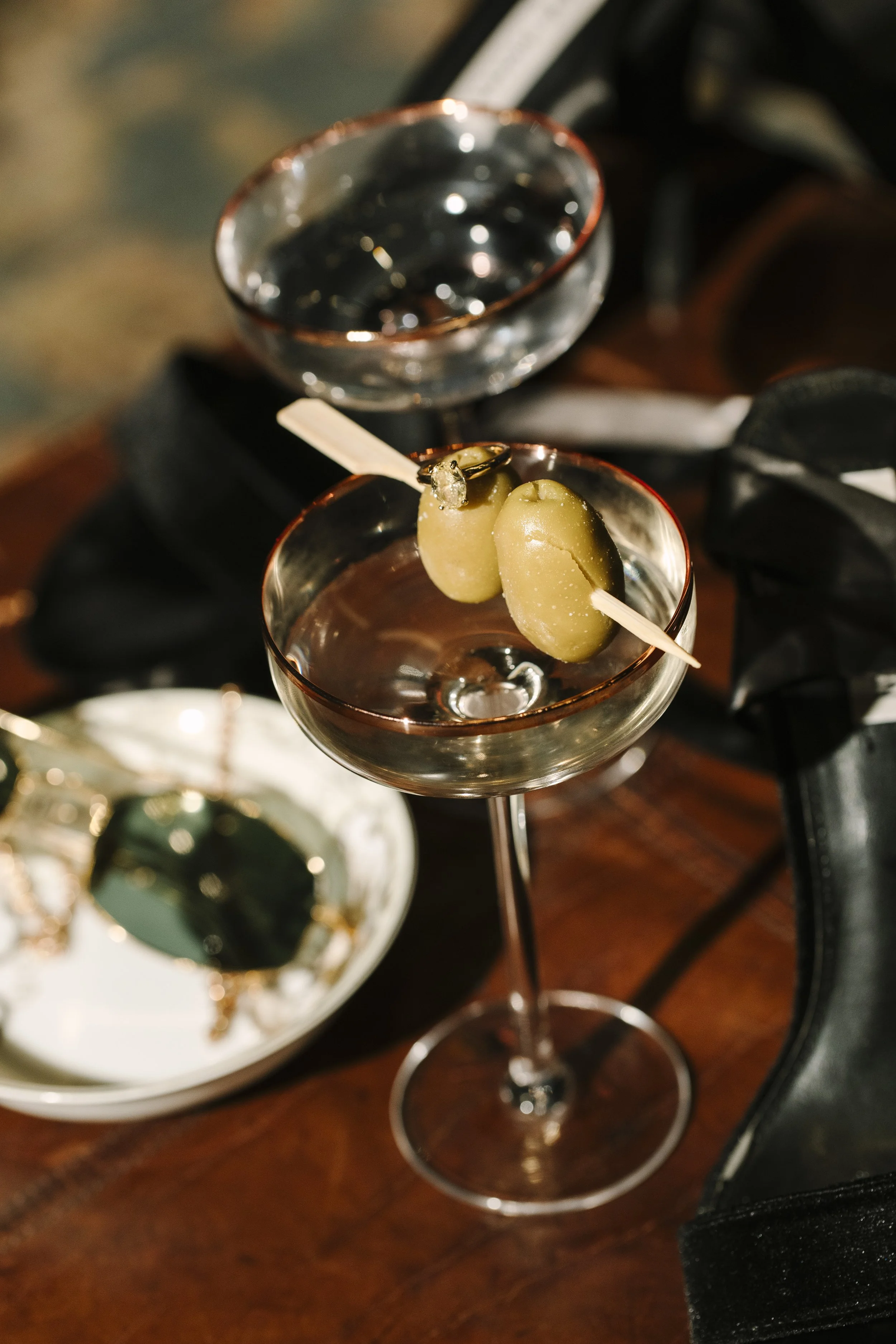 Close-up of a cocktail garnished with green olives on a pick, surrounded by drinks on a dimly lit bar table.