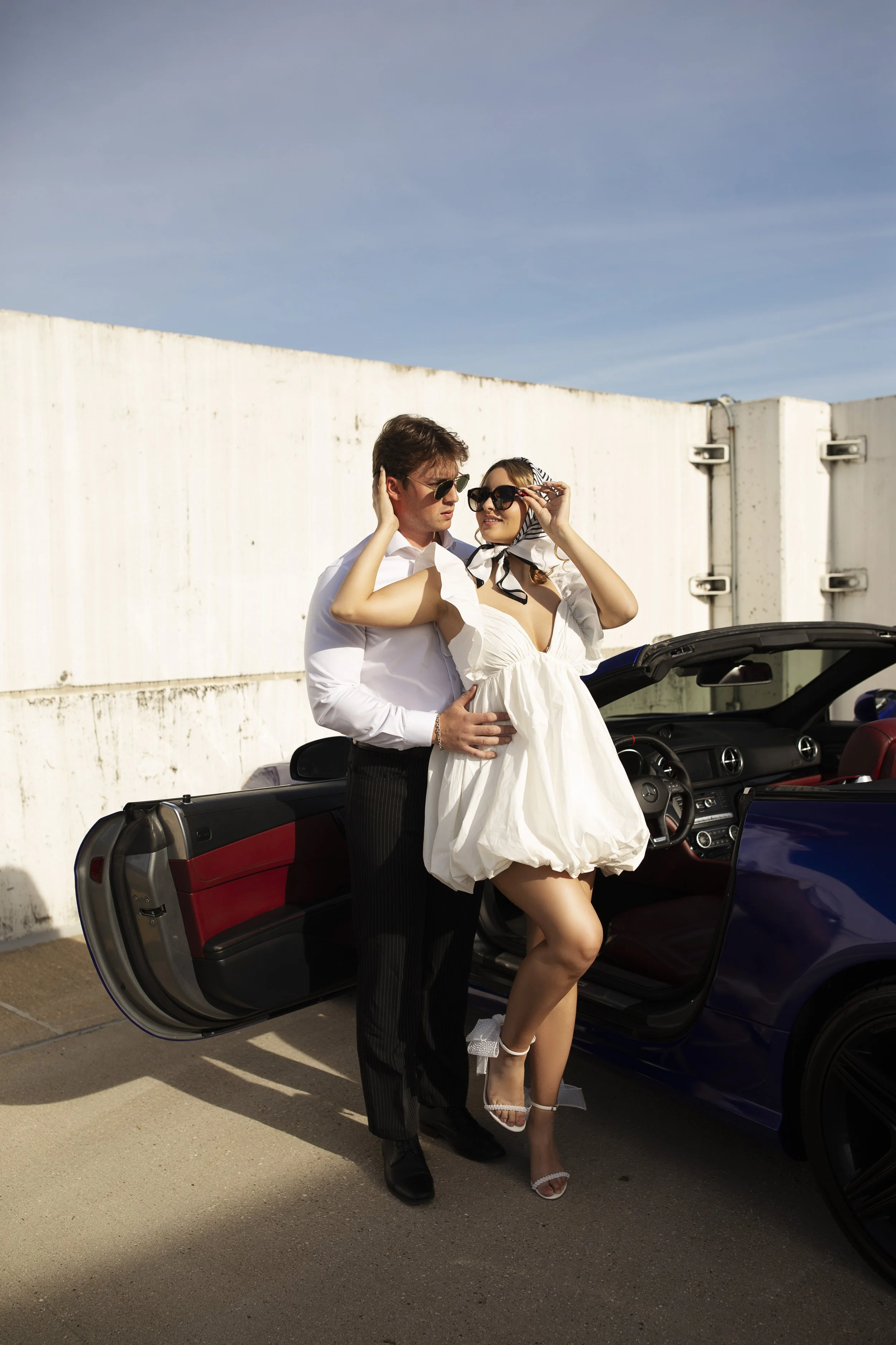 Stylish couple wearing sunglasses beside an open convertible car on a sunny rooftop