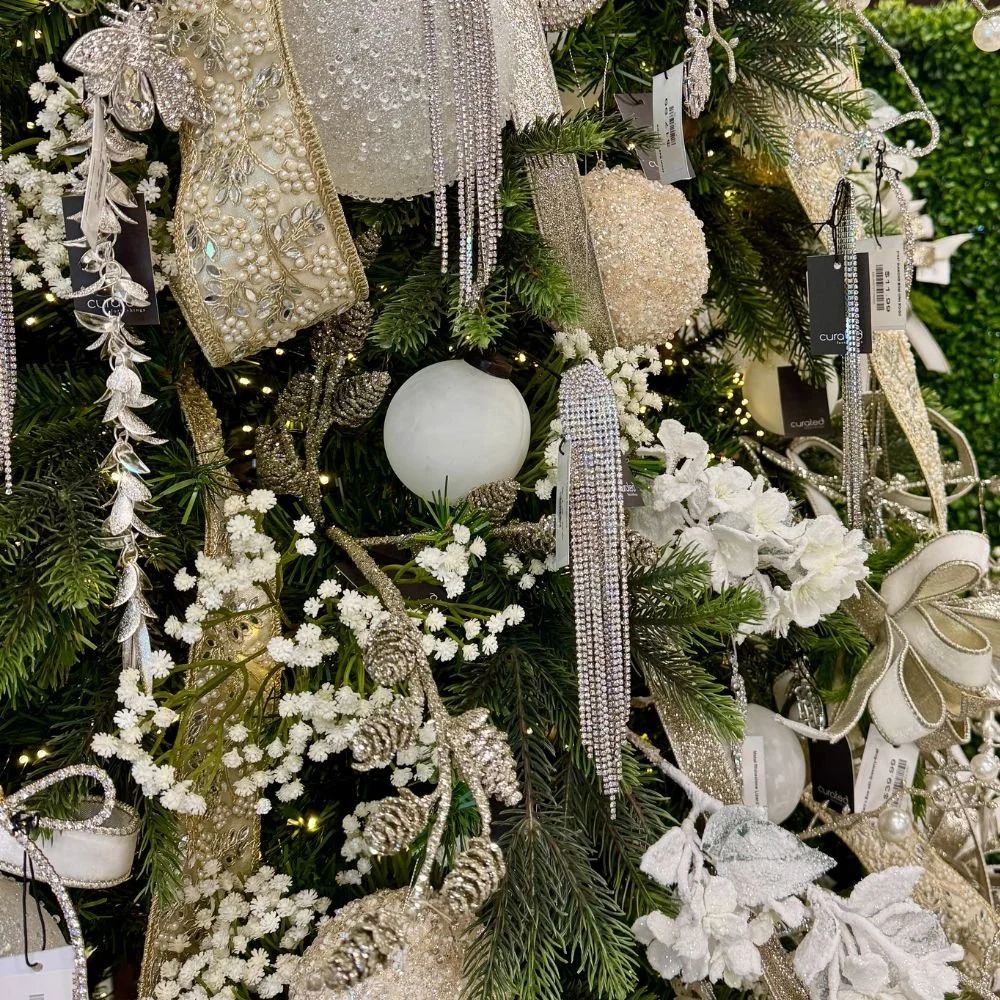 White Christmas ornaments and soft velvet ribbon styled among deep evergreen branches with sparkling accents.