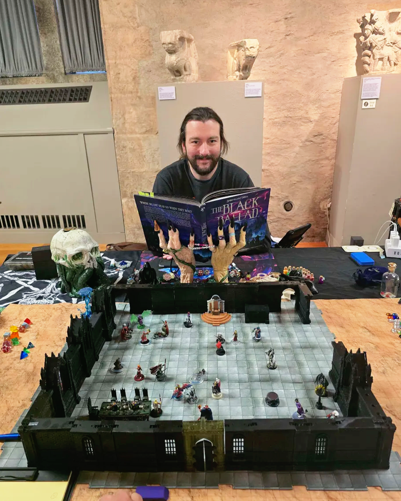 A man with long hair and a beard sitting behind a table playing Dungeons & Dragons, with a gaming set, dice, miniatures, and accessories on the table, and a large fantasy-themed book titled "The Black Ballad" open in front of him.