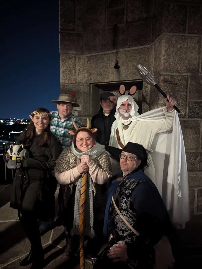 Six people in costumes posing against a stone wall and night sky. One in a wizard outfit with a staff, another with deer antlers, a woman with fairy wings, a woman with a Viking outfit, a man with glasses and a hat, and a man in a plaid shirt and hat.