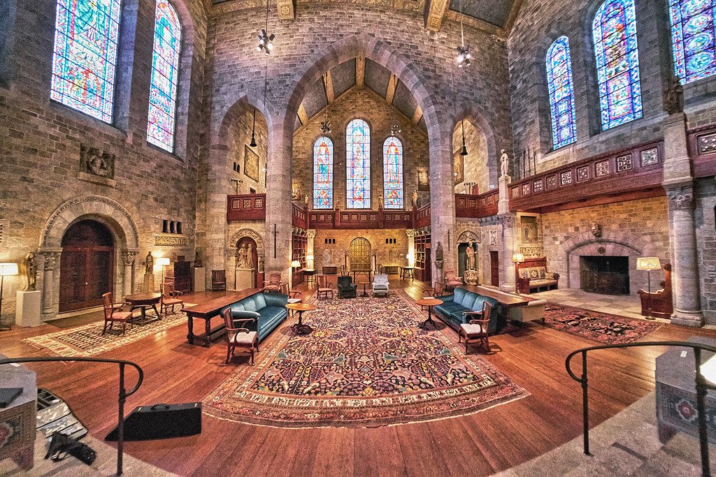 A group of people gathered around a table playing a board game in a room with wooden floors and stone walls, decorated with religious sculptures and statues, illuminated by lamps.