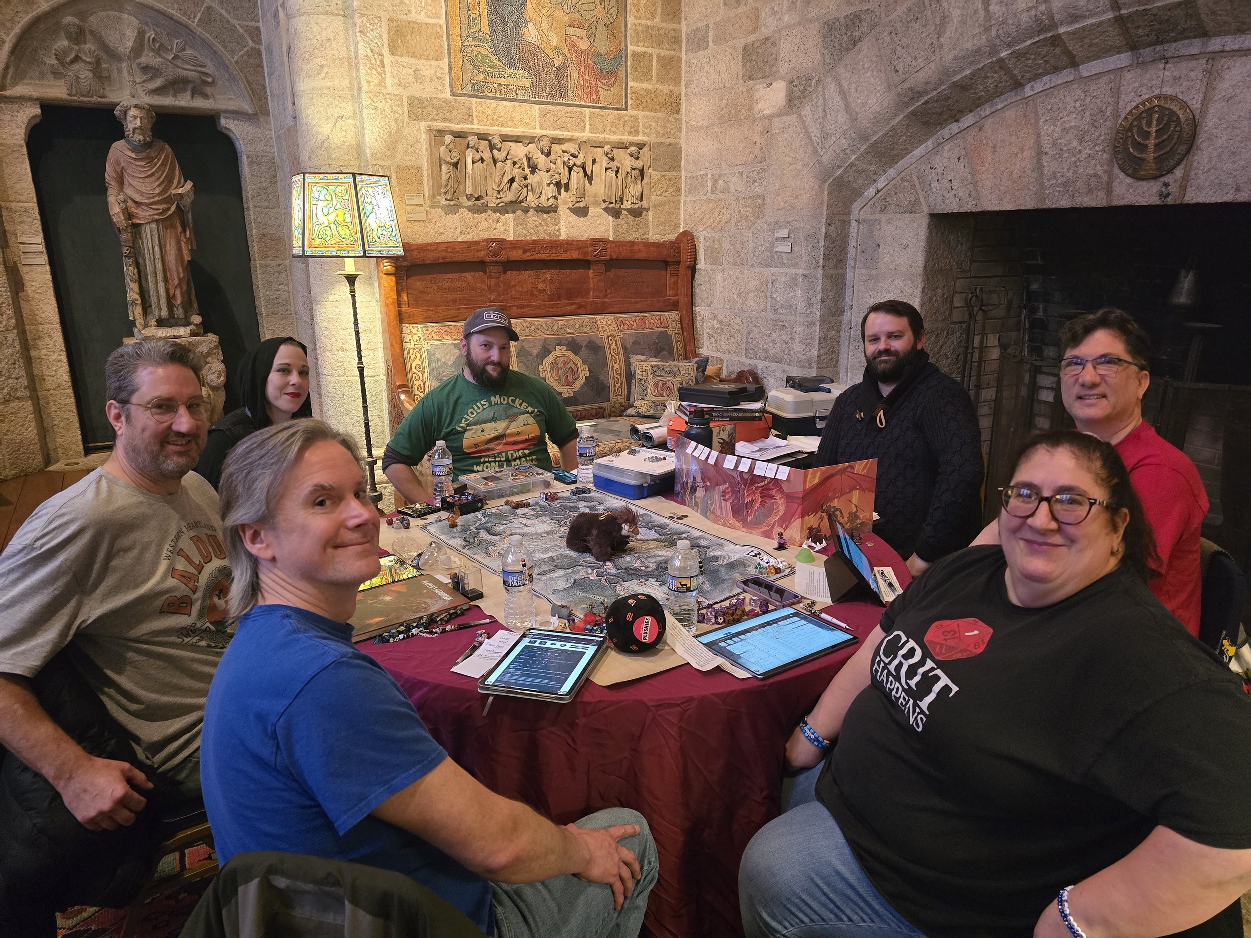 A group of people around a gaming table in a room with stone walls and statues. One person is standing, pointing, while others are seated, some using laptops or tablets. The table has gaming accessories, miniatures, and drinks.