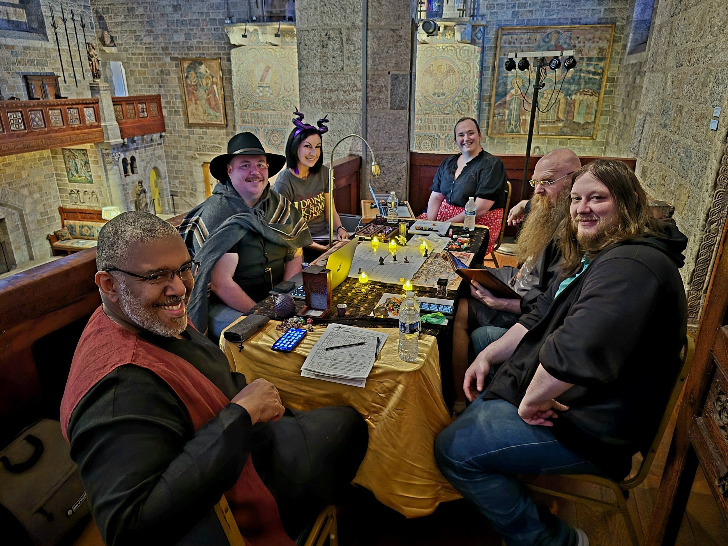 A group of people around a gaming table in a room with stone walls and statues. One person is standing, pointing, while others are seated, some using laptops or tablets. The table has gaming accessories, miniatures, and drinks.