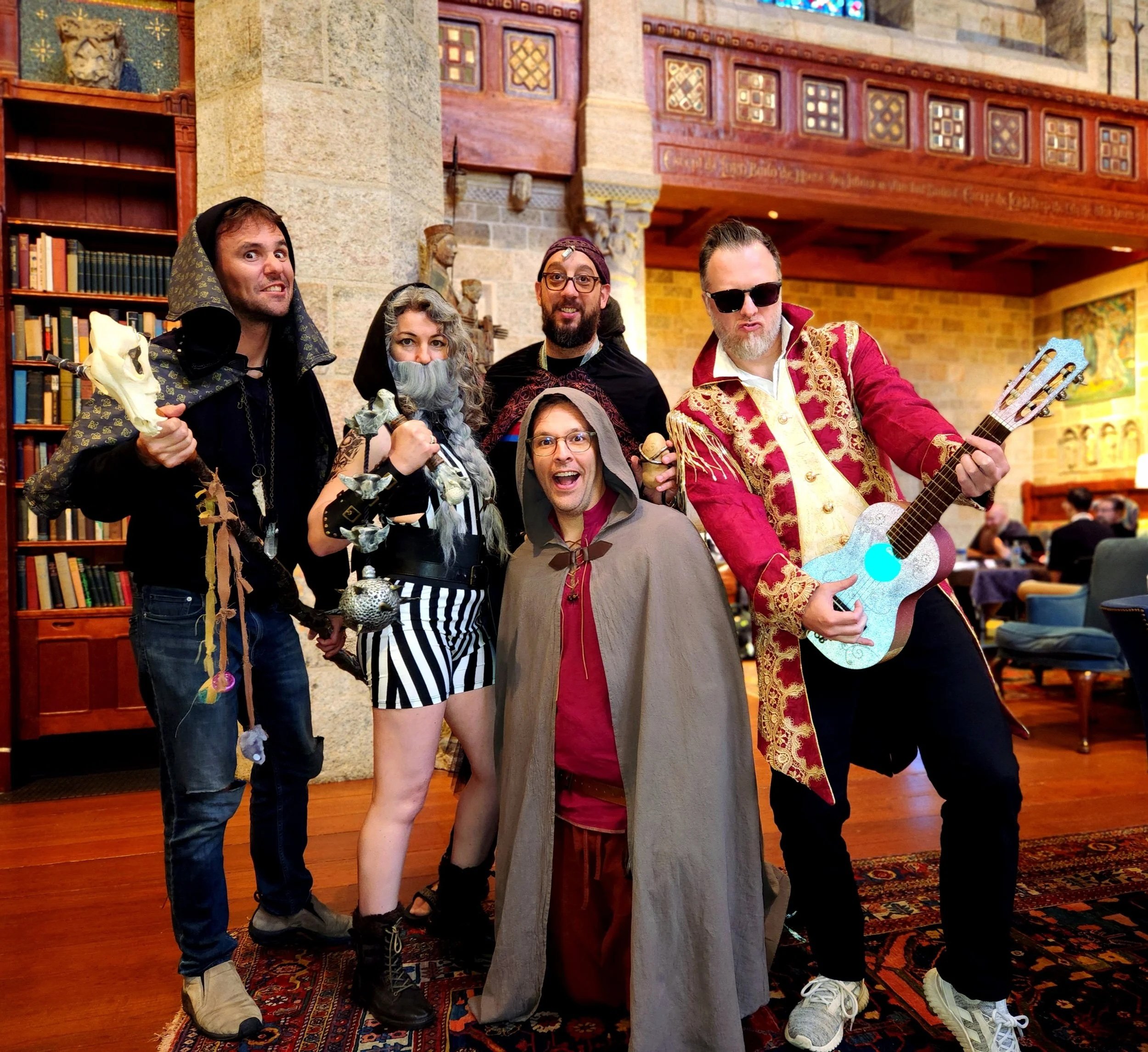 Six people in costumes posing against a stone wall and night sky. One in a wizard outfit with a staff, another with deer antlers, a woman with fairy wings, a woman with a Viking outfit, a man with glasses and a hat, and a man in a plaid shirt and hat.