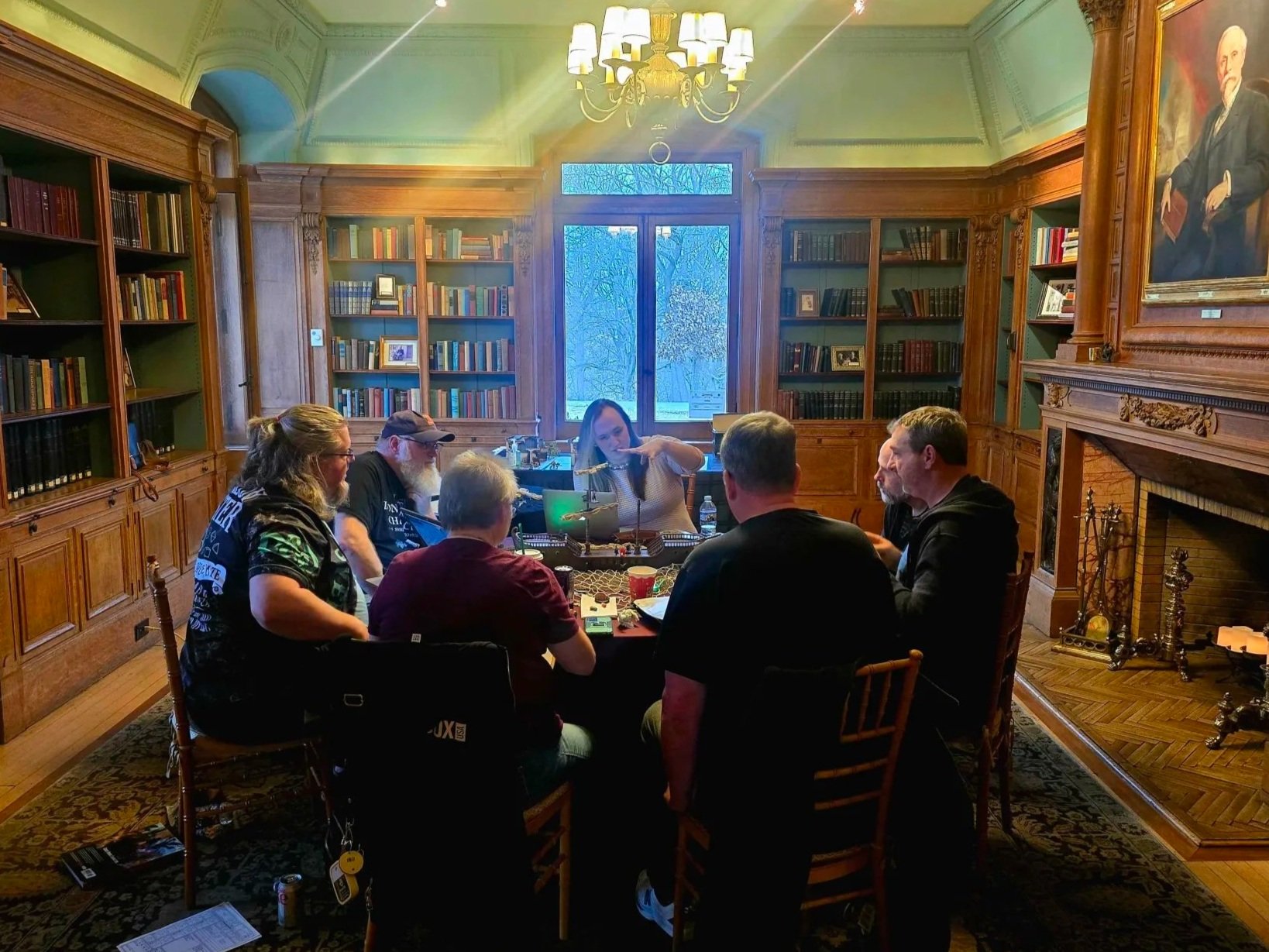 A group of people around a gaming table in a room with stone walls and statues. One person is standing, pointing, while others are seated, some using laptops or tablets. The table has gaming accessories, miniatures, and drinks.