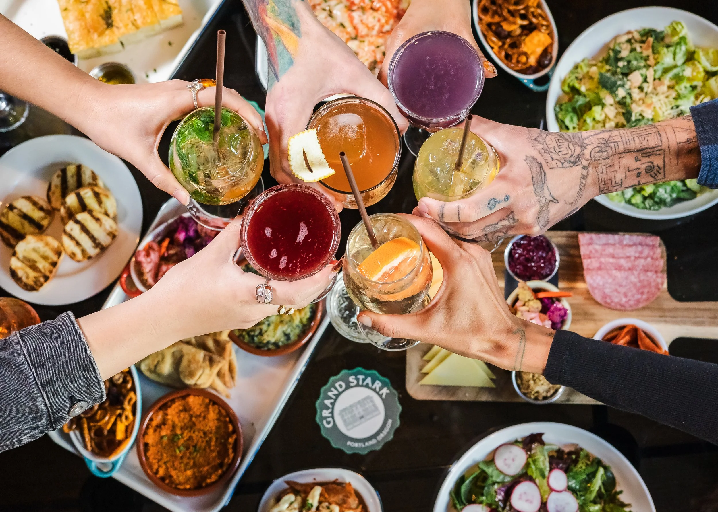 Overhead view of friends clinking colorful cocktails over a table filled with assorted food