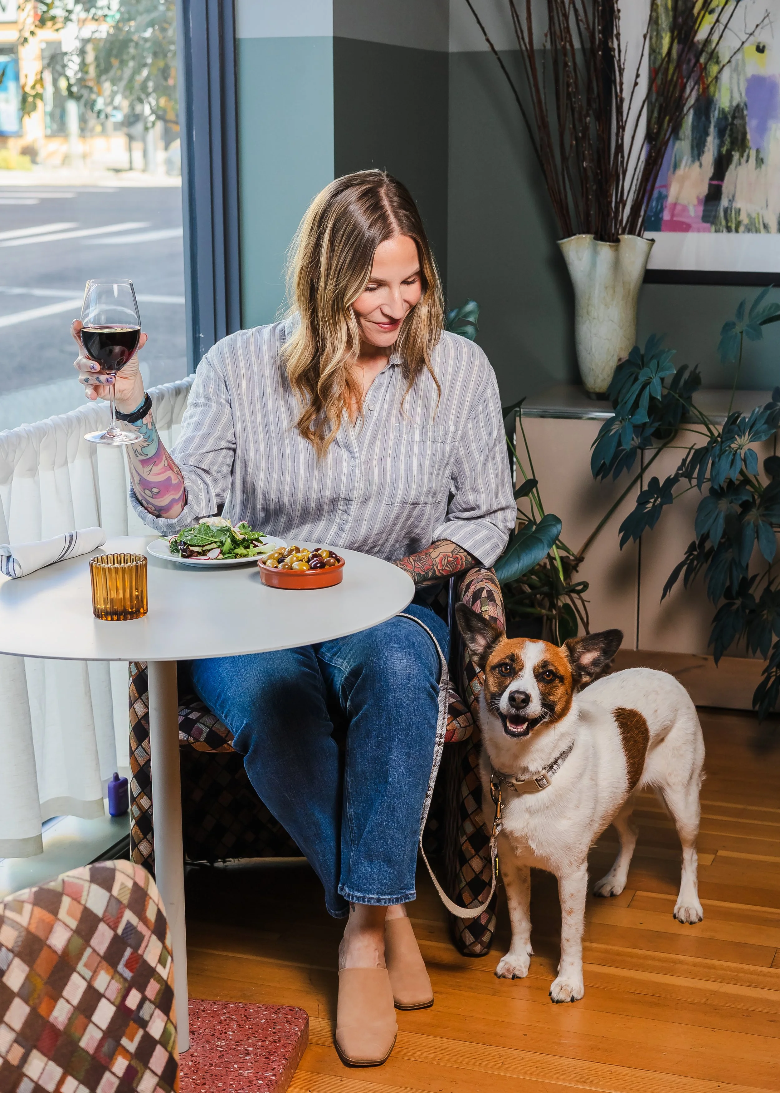Tattooed woman dining at a café table with a glass of red wine, salad, olives, and her dog beside her.