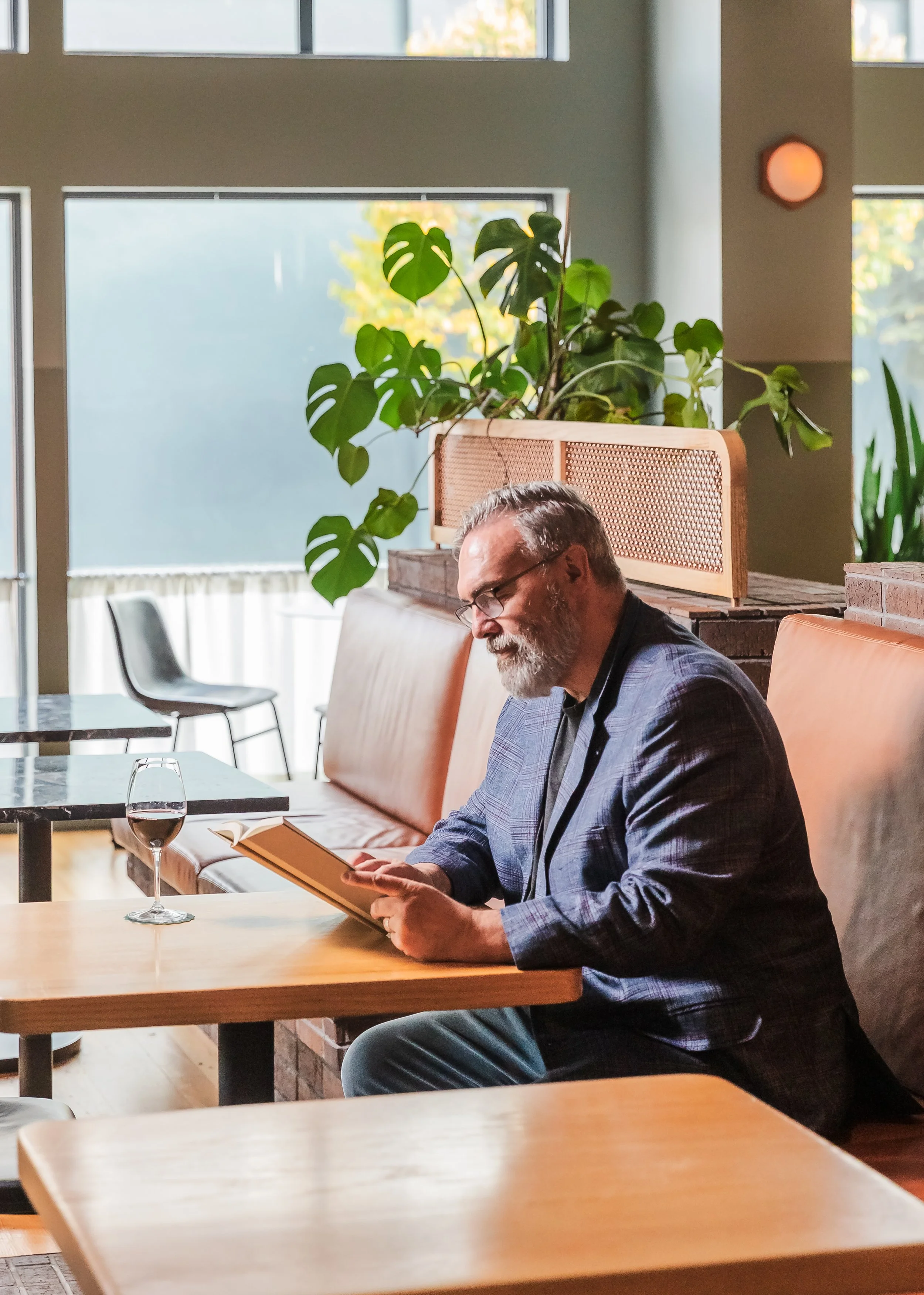 Bearded man reading a book at a bar table with a glass of red wine and indoor plants behind him