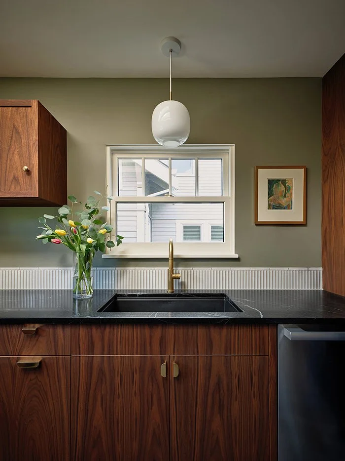 A brass kitchen sink centered in front of a window. The walls are green and the cabinetry is walnut. 