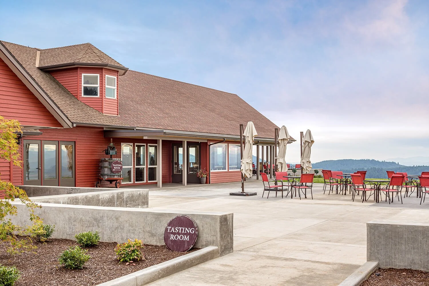 A red barn houses a tasting room with a large concrete patio. Rolling hills are visible in the distance.