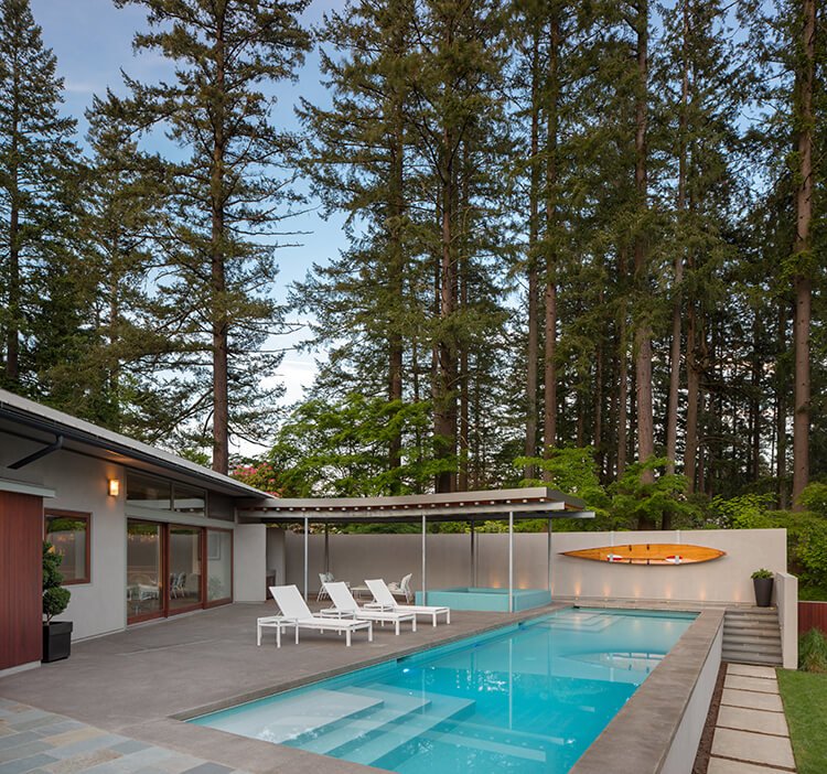 A swimming pool at a custom built residential home. The water is clear blue, white lounge chairs and an orange kayak line the sides. There are tall fir trees in the background.

