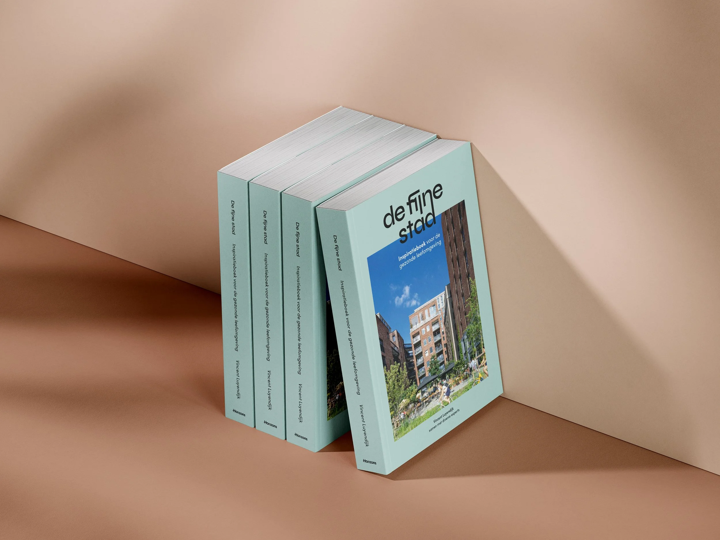 Vijf boeken met de titel 'de fijne stad' staan tegen een beige muur. De boeken hebben een lichtblauwe omslag met een foto van een stedelijk parkertje en moderne gebouwen.