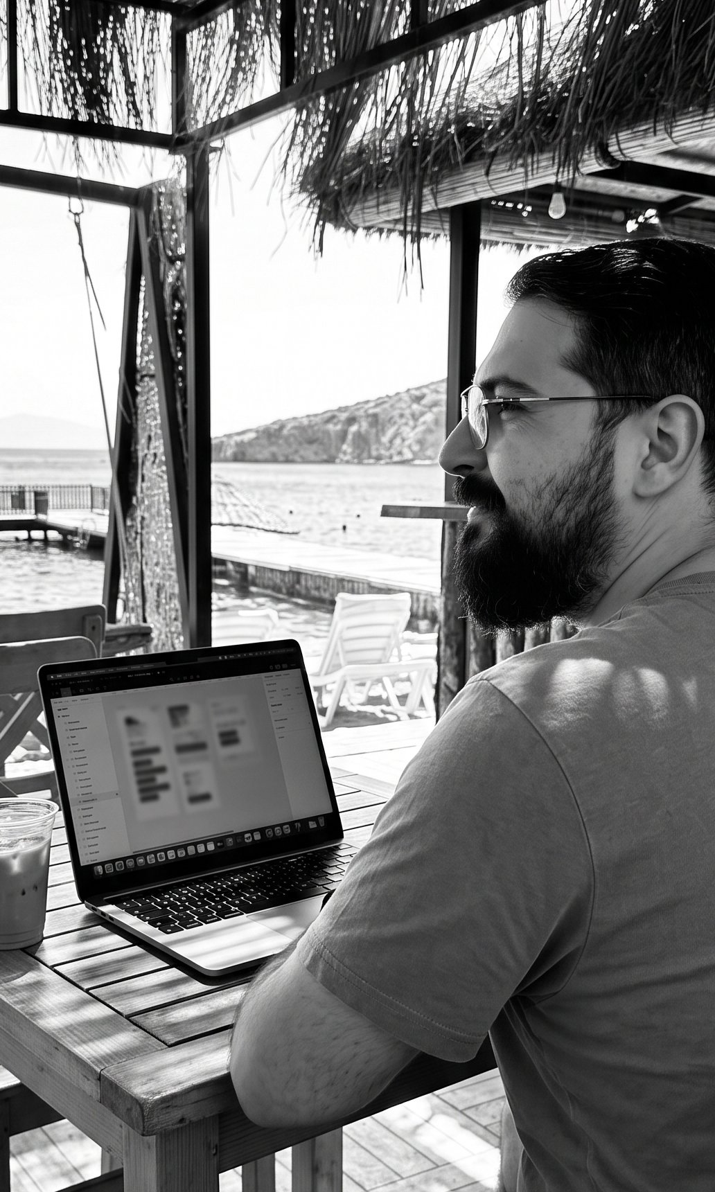 A black and white photo of a man with glasses and a beard working on a laptop at an outdoor cafe near water, with boats and a mountain in the background.
