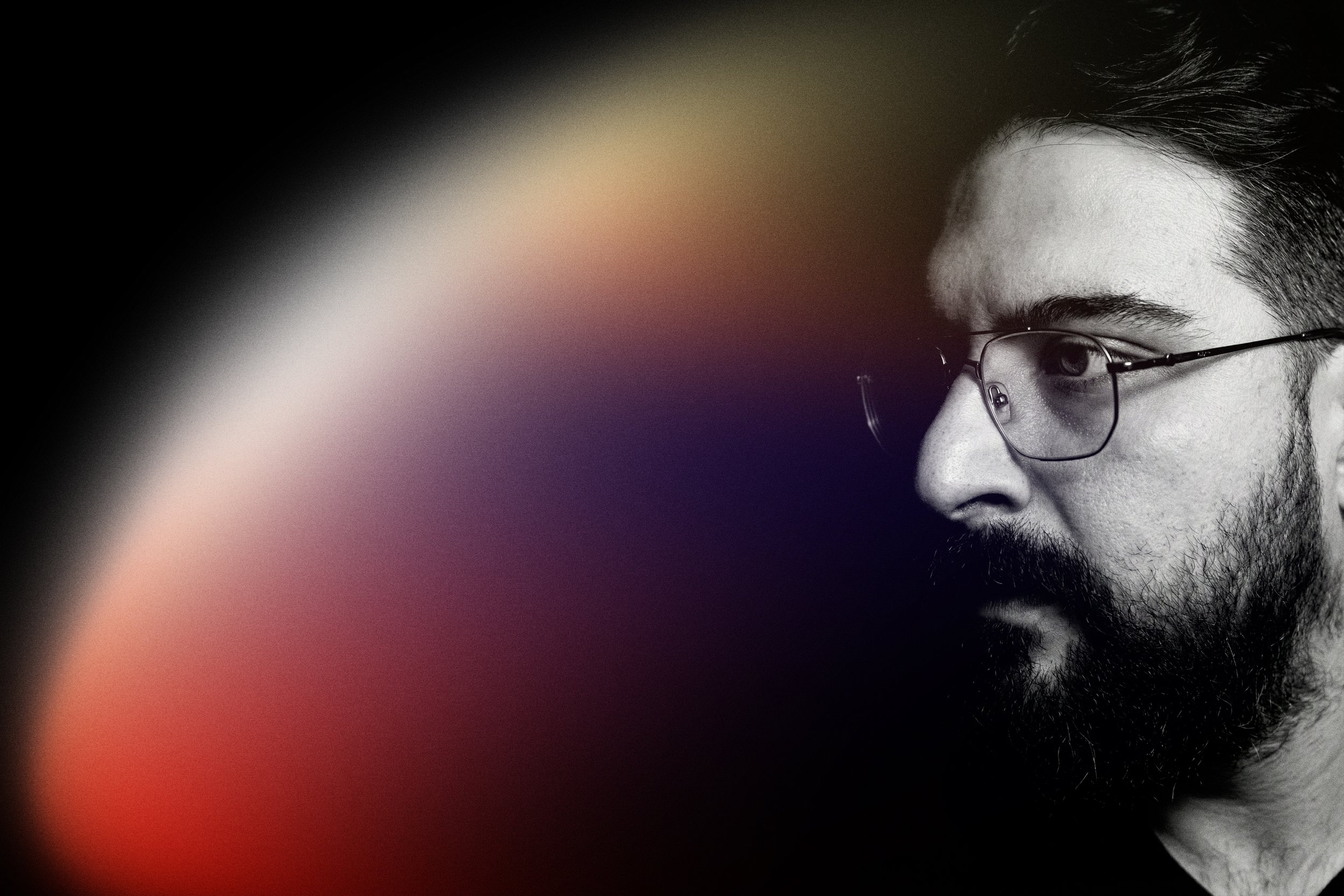 Close-up of a man with glasses and a beard, looking to the right, with a blurred multicolored background.