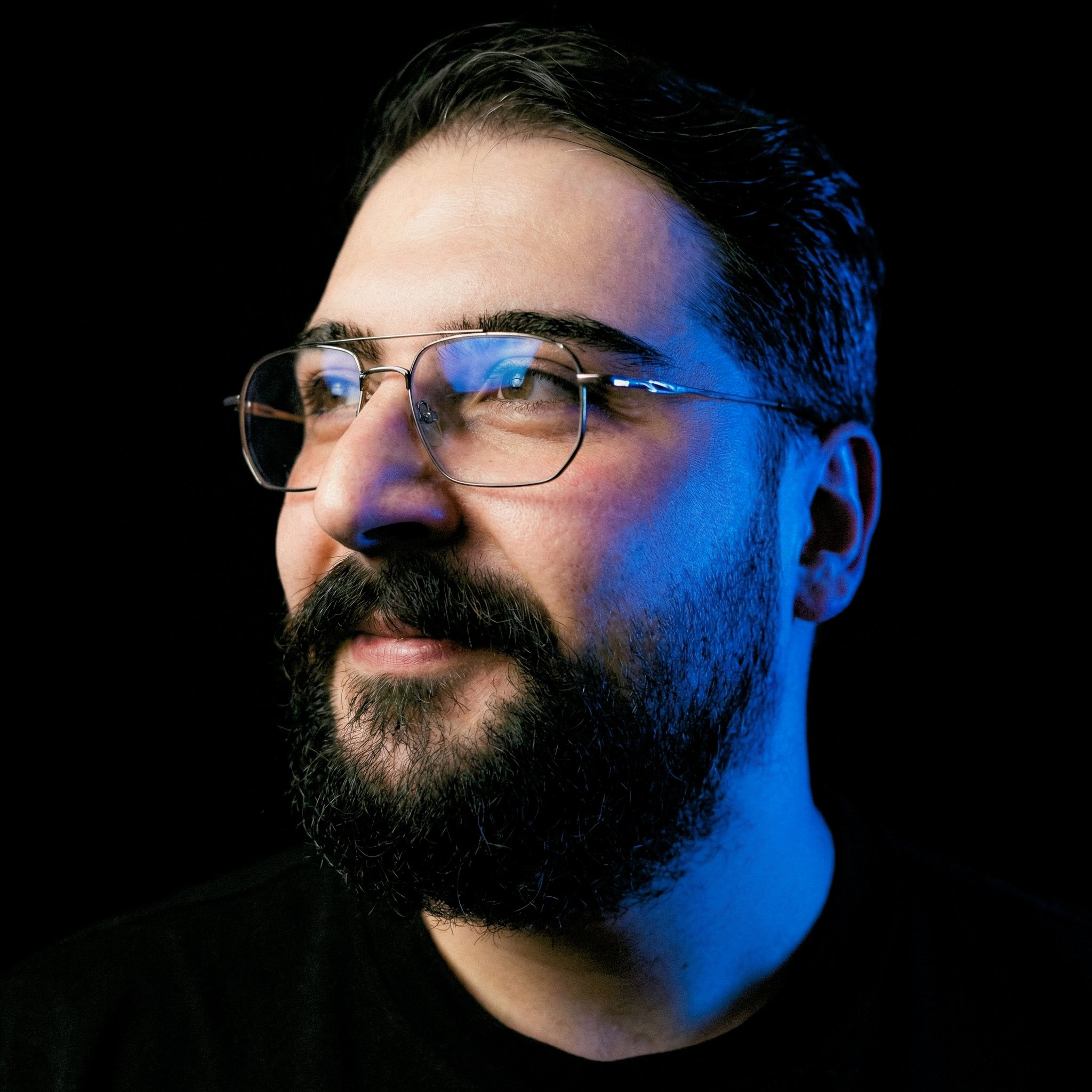Oğuz Atılan's photo, close-up of a man's face with glasses, dark hair, and a beard, illuminated with blue lighting on the right side, against a black background.