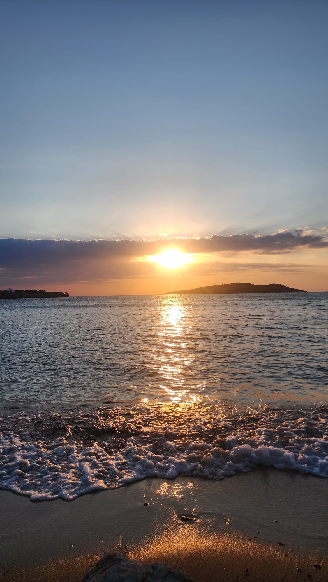 A sunset over the ocean with the sun near the horizon, reflecting on the water, with small waves and a sandy beach in the foreground.