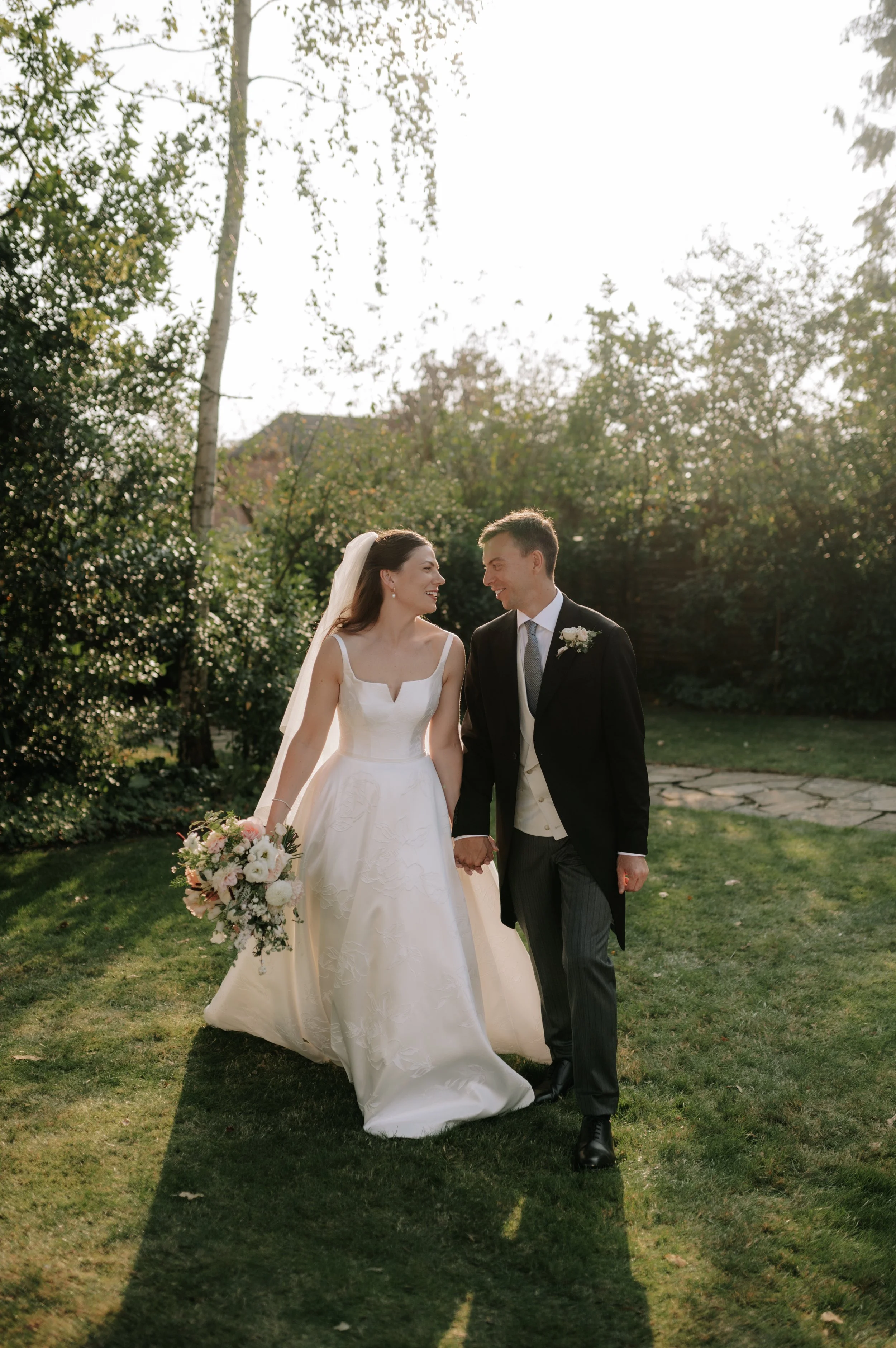 bride and groom walk holding hands smiling at each other. bride wearing Suzanne neville.