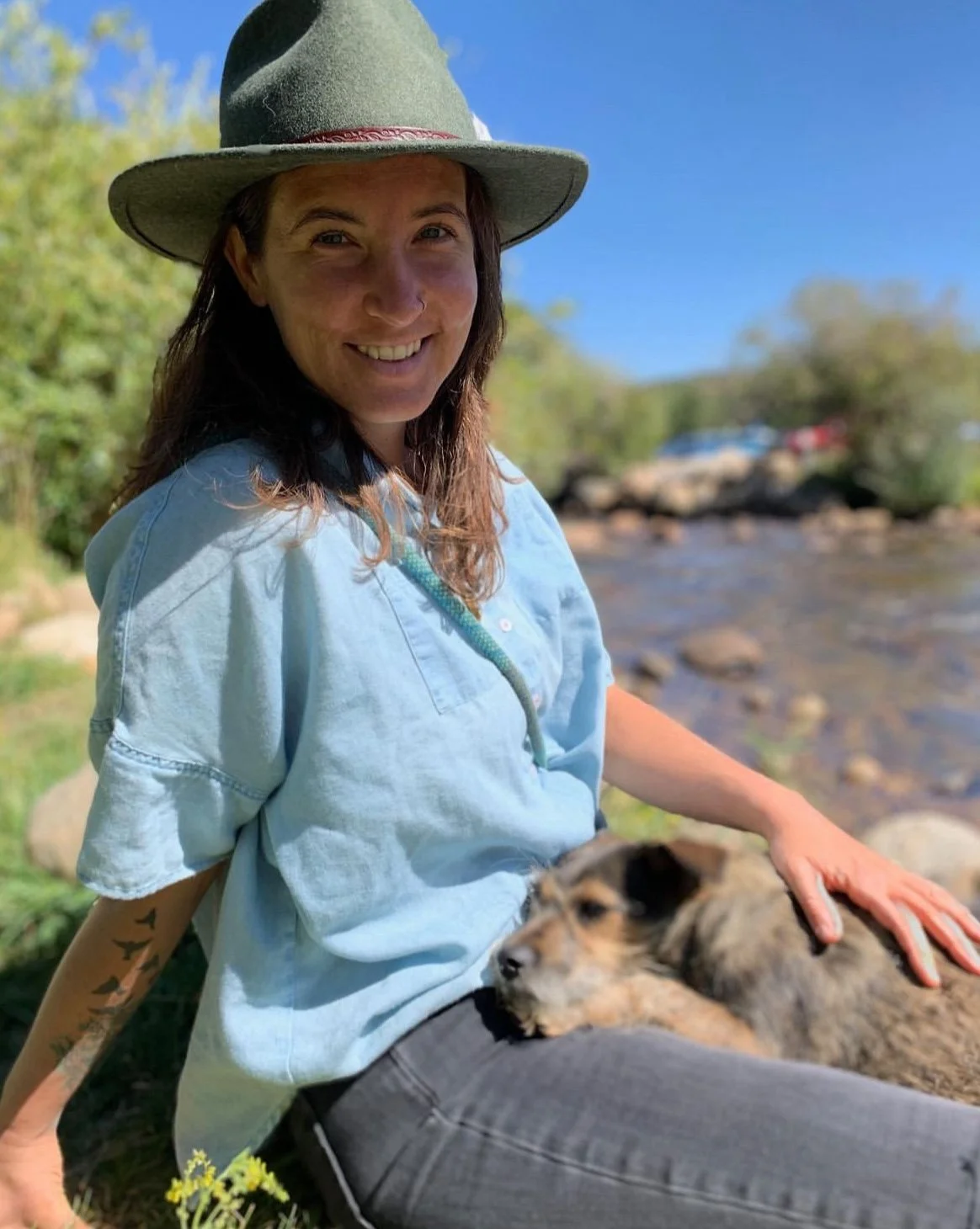 Person wearing a green hat and blue shirt sits by a river with a small dog on their lap.