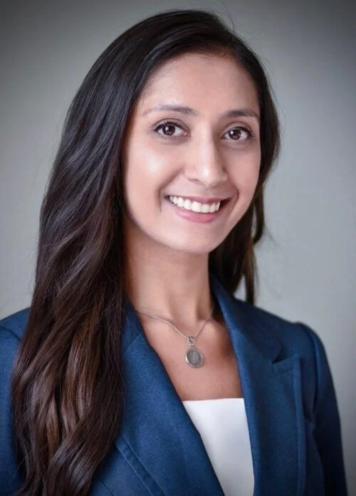 Portrait of a woman with long dark hair, wearing a blue blazer and a necklace, smiling at the camera.