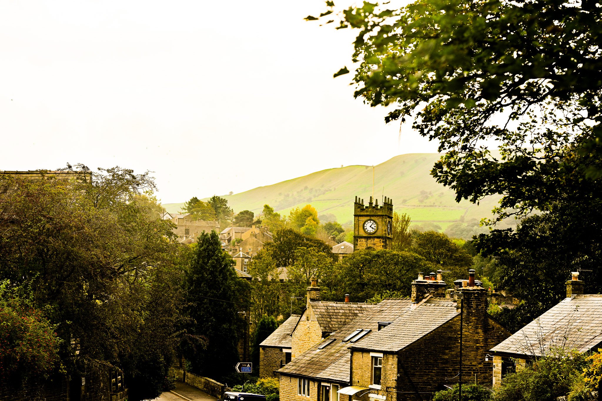 A small town with stone houses and a clock tower, green trees, and hills in the background.