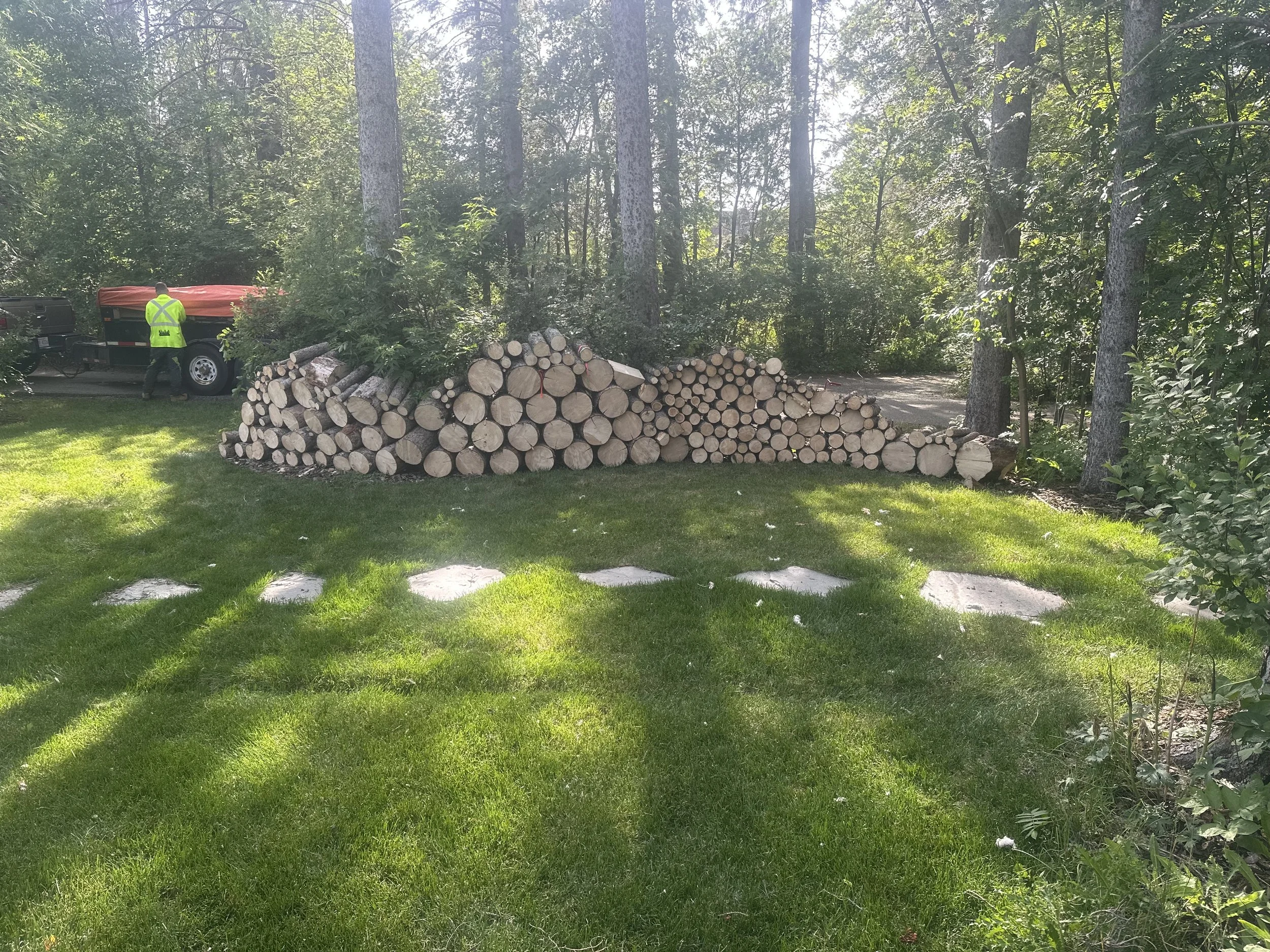 A stack of cut logs is arranged in a garden with green grass and trees, with a person in a yellow safety vest near a trailer in the background.