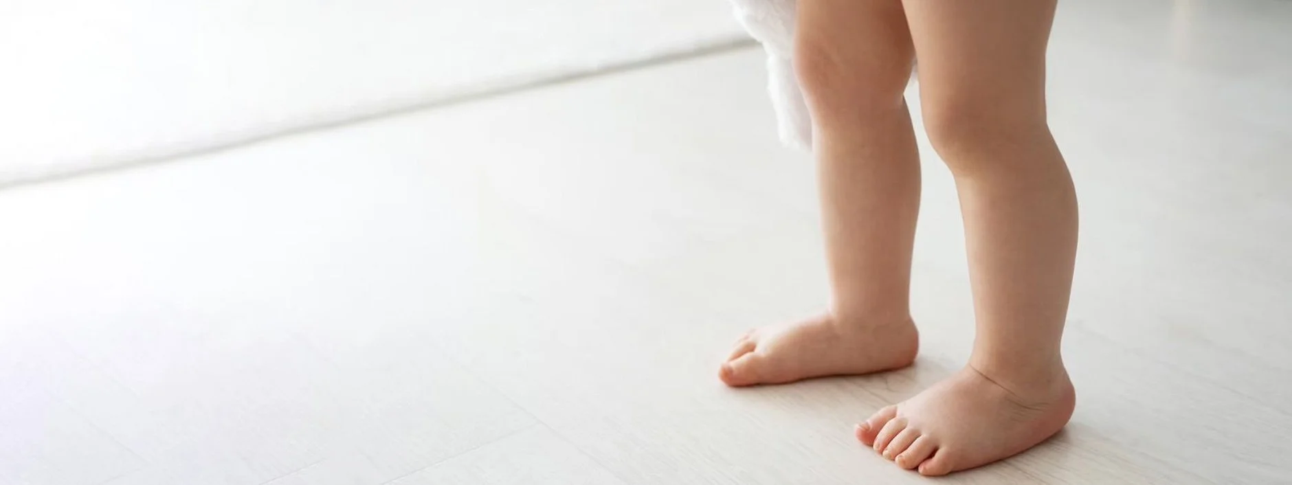 Pied d'un bébé debout sur un sol en bois clair dans une pièce lumineuse.