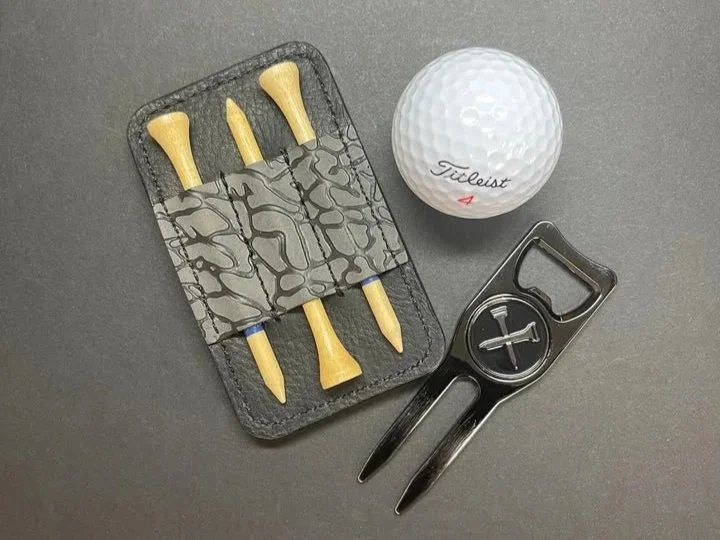 A set of golf tee markers in a black case, a Titleist golf ball, and a golf ball marker with a divot repair tool on a gray surface.