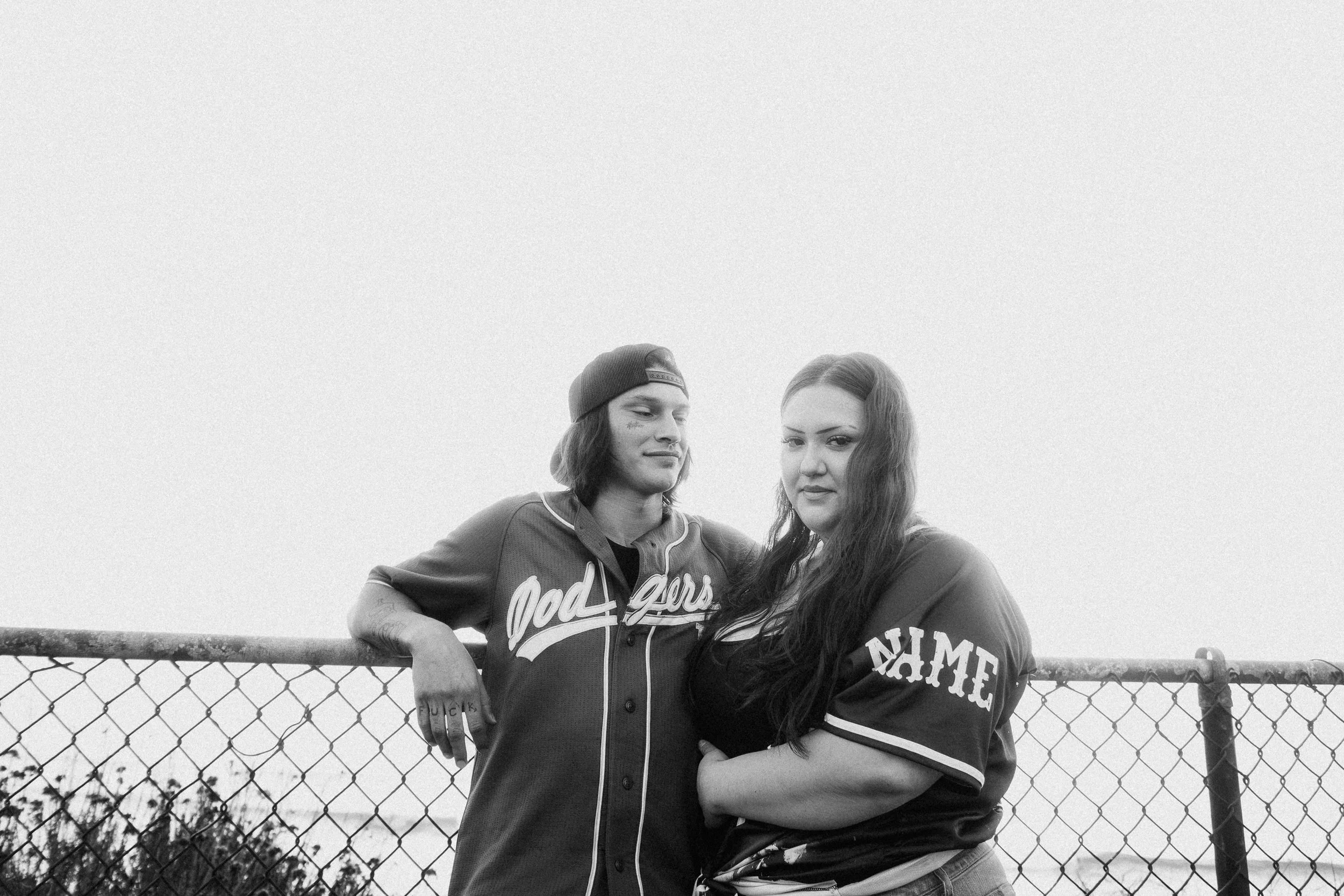 Two women standing close together behind a chain-link fence outdoors, with a cloudy sky in the background.