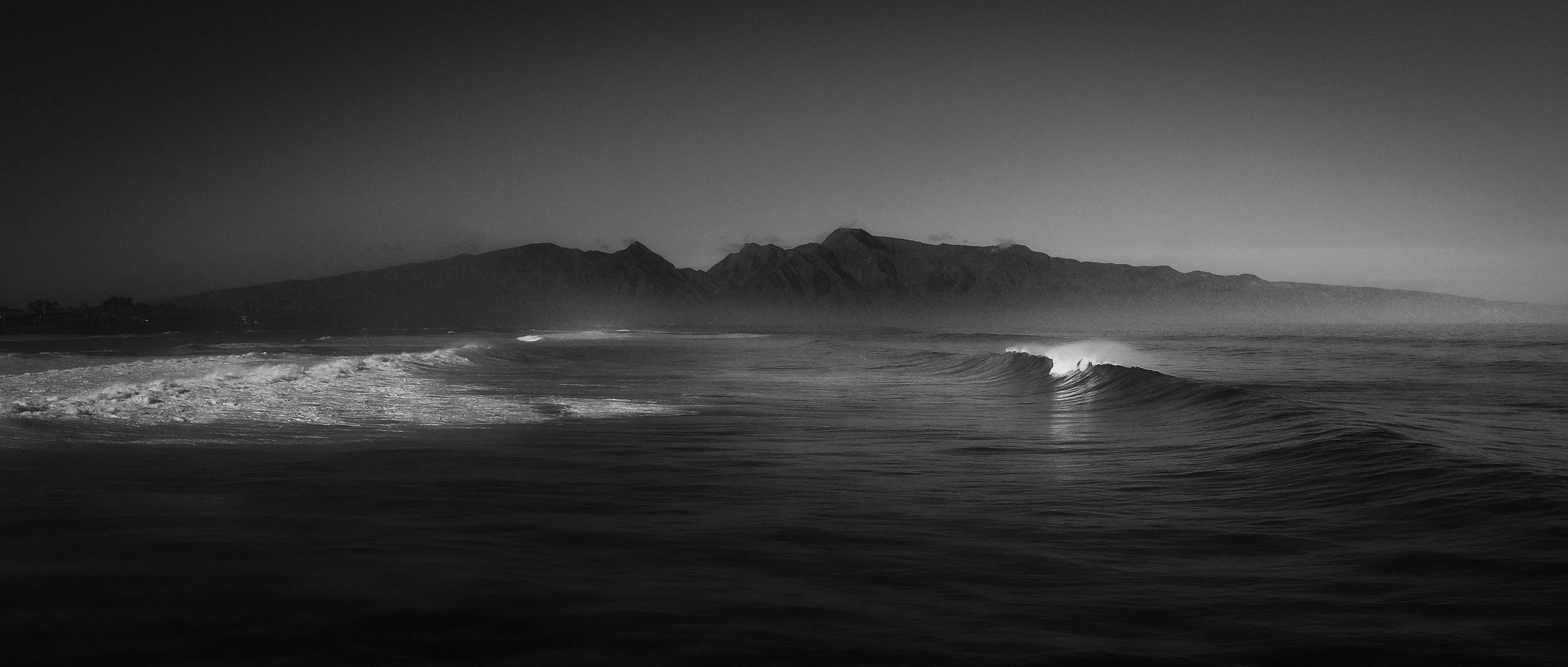 Black and white photograph of ocean waves approaching the shoreline with mountainous terrain in the background.