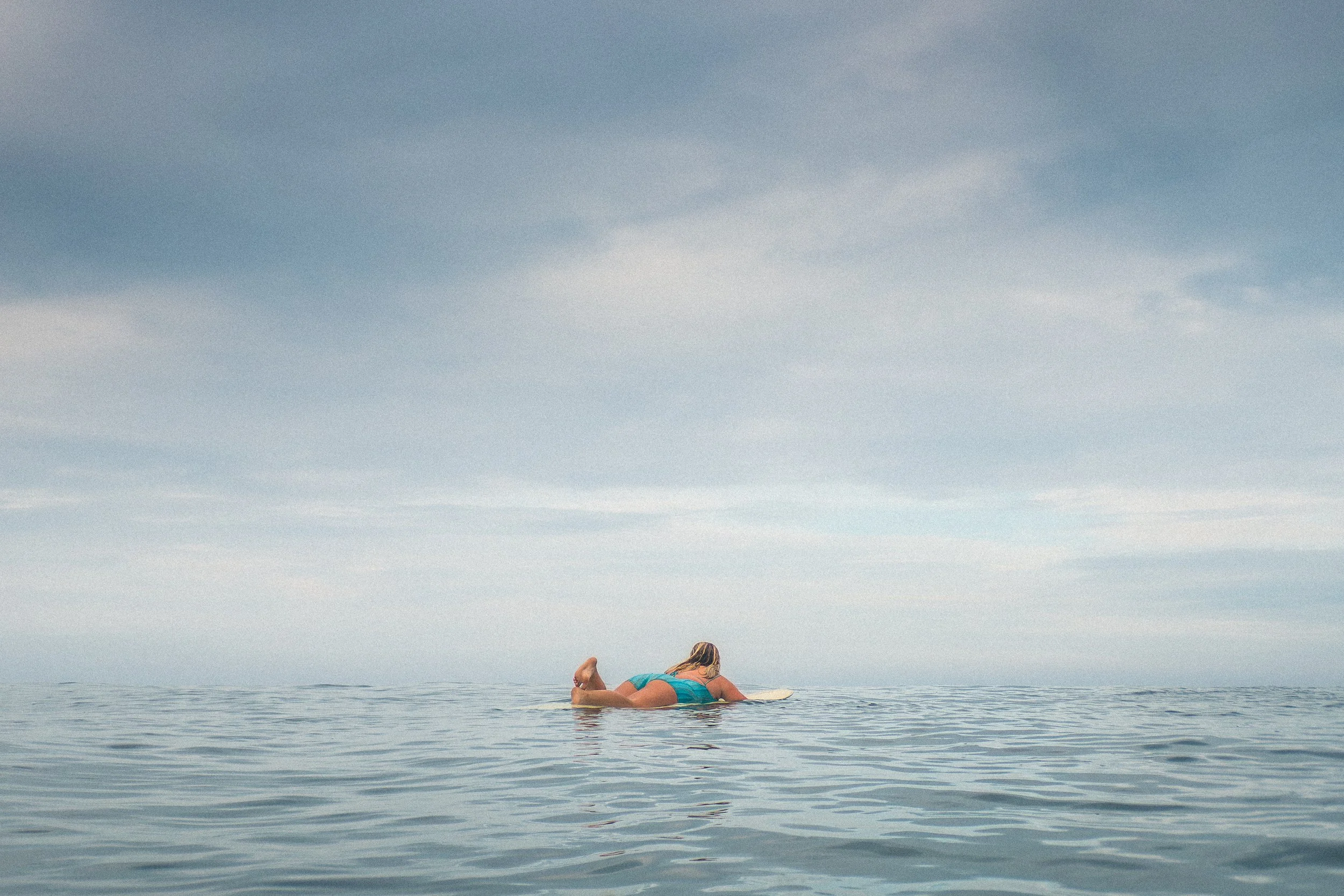A woman in a turquoise swimsuit relaxing on a surfboard in calm ocean water, with a vast cloudy sky overhead.
