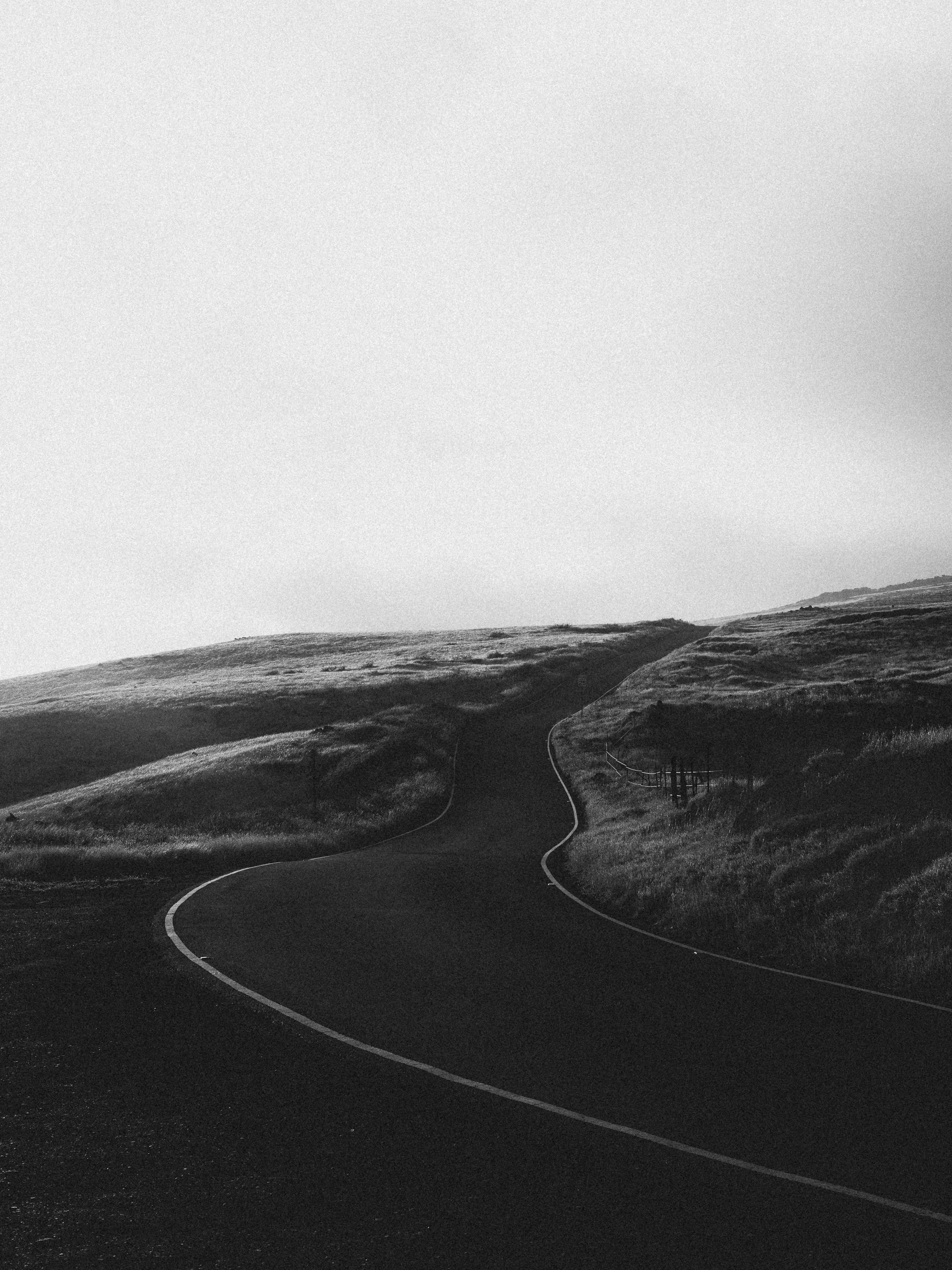 A winding road in a hilly landscape with grassy terrain, fencing, and hills in the background under a clear sky.