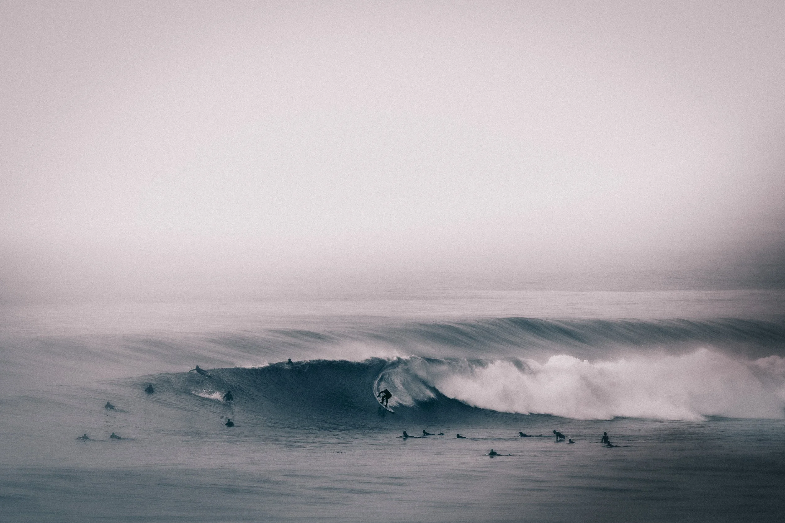 Multiple surfers riding and waiting for waves in the ocean, overcast sky, in a black-and-white tone.
