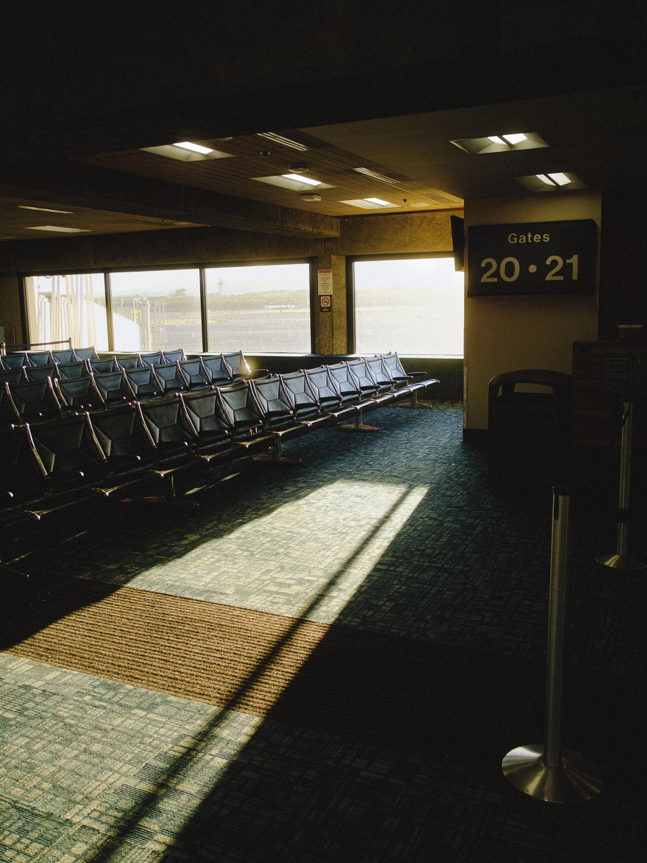 Empty airport gate area with rows of black seats, sunlight coming through large windows, and a sign indicating gates 20 and 21.