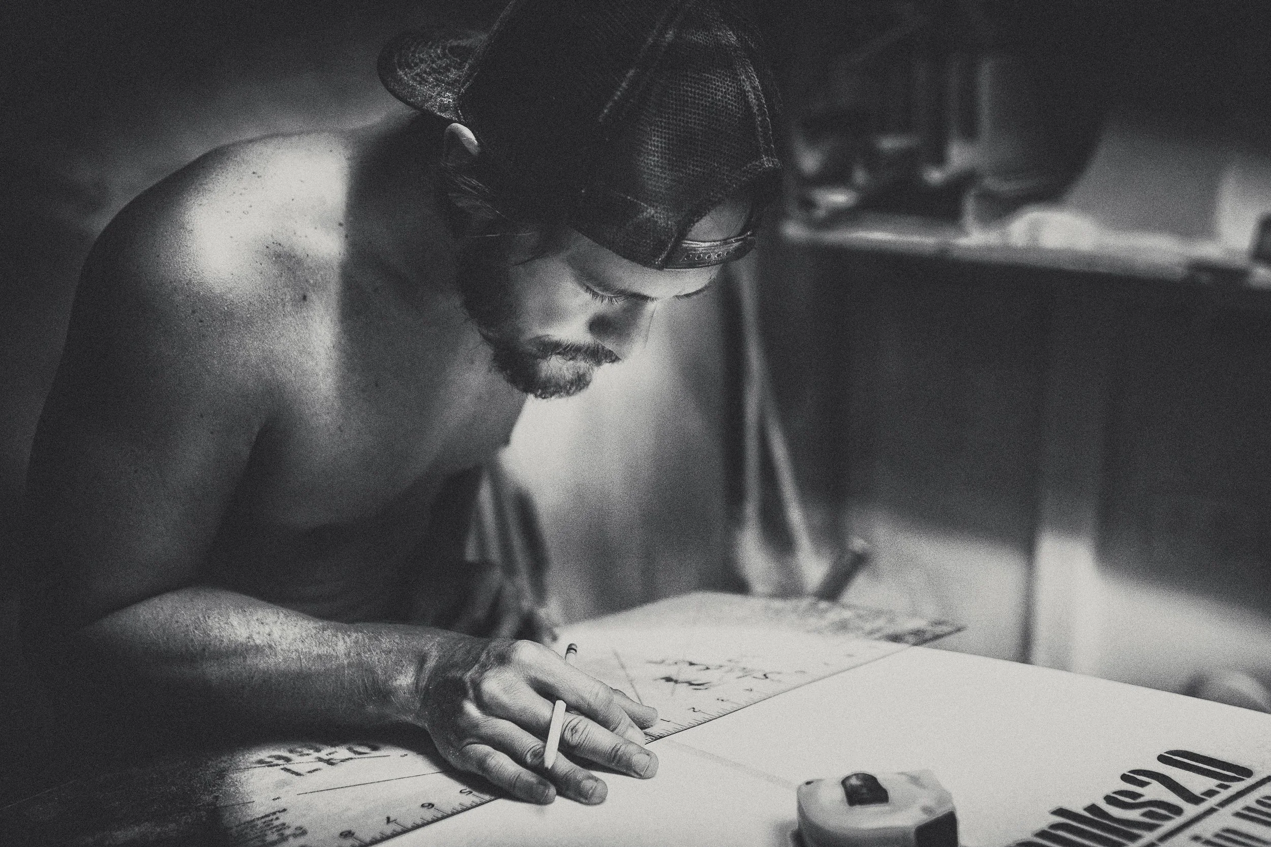 A man with a beard and cap drawing or writing on a large sheet of paper with a ruler. The scene is in black and white.