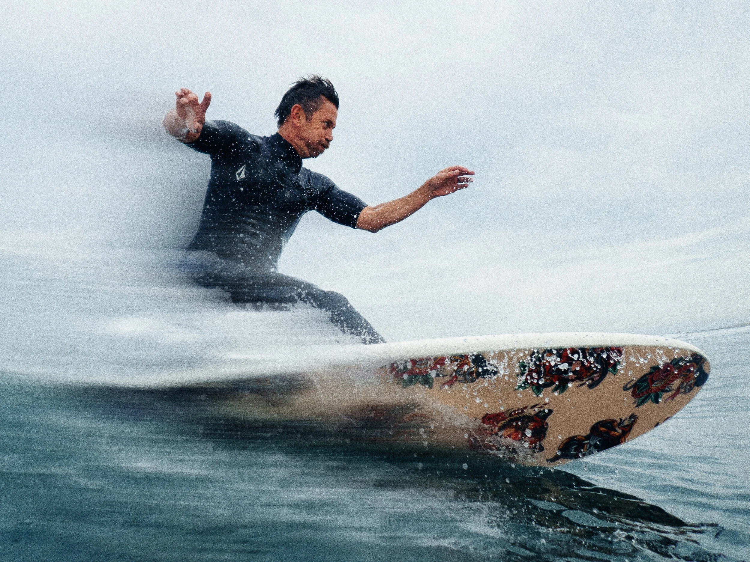 Man sitting on surfboard in the ocean, with a tattooed leg visible, water splashing around him, overcast sky in the background.