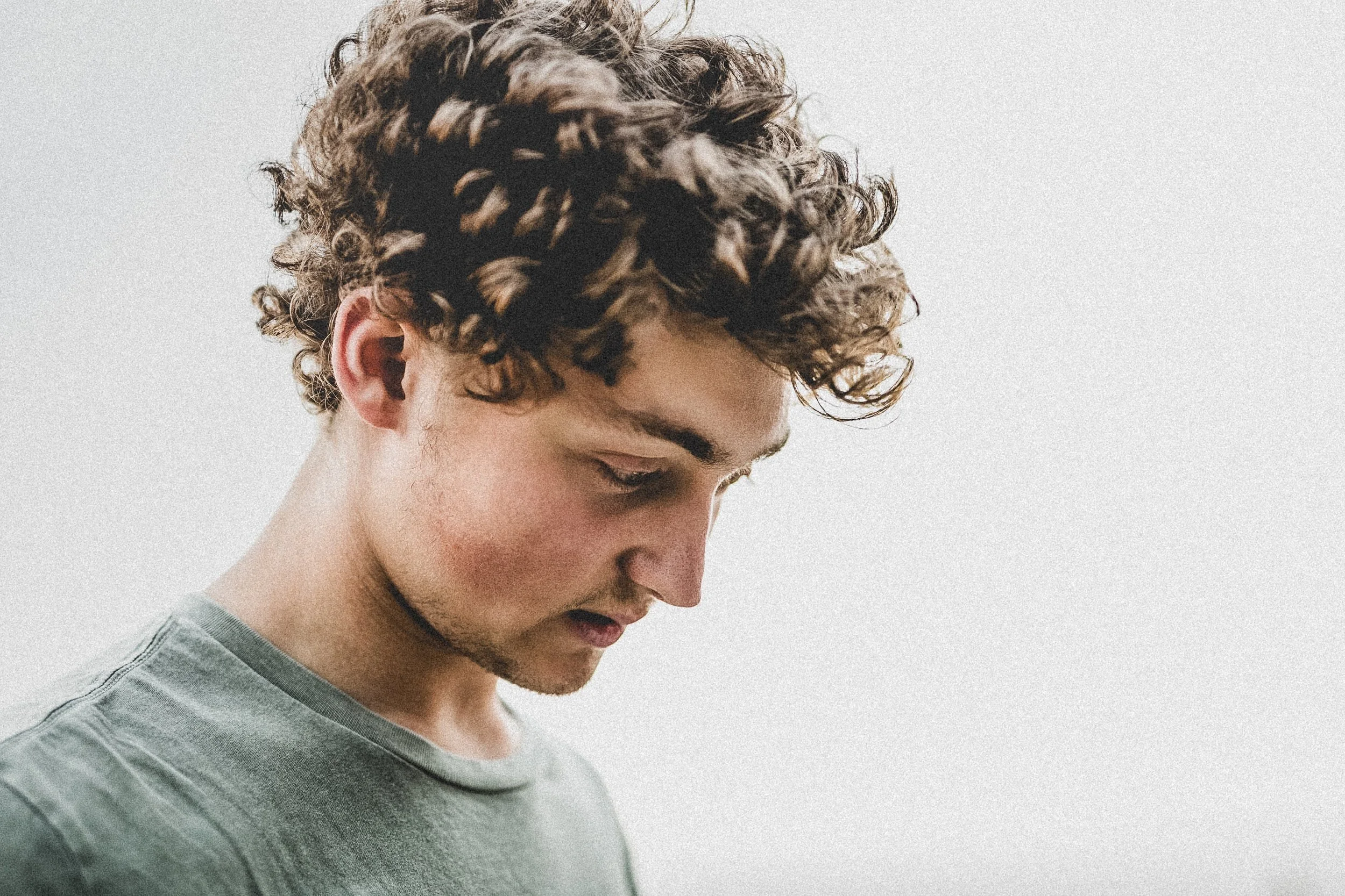 Side profile of a young man with curly brown hair and light skin, wearing a light gray shirt, looking downward against a plain light background.