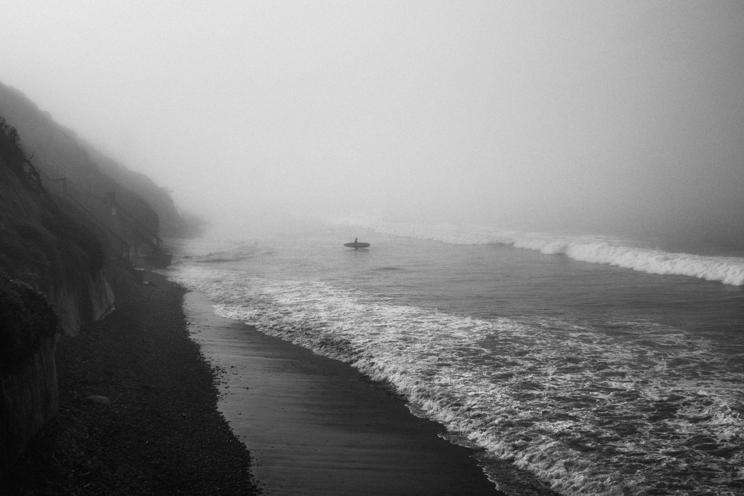 A foggy beach scene with dark shoreline, small waves, a lone surfer with a surfboard, facing the ocean, and overhanging cliffs on the left.