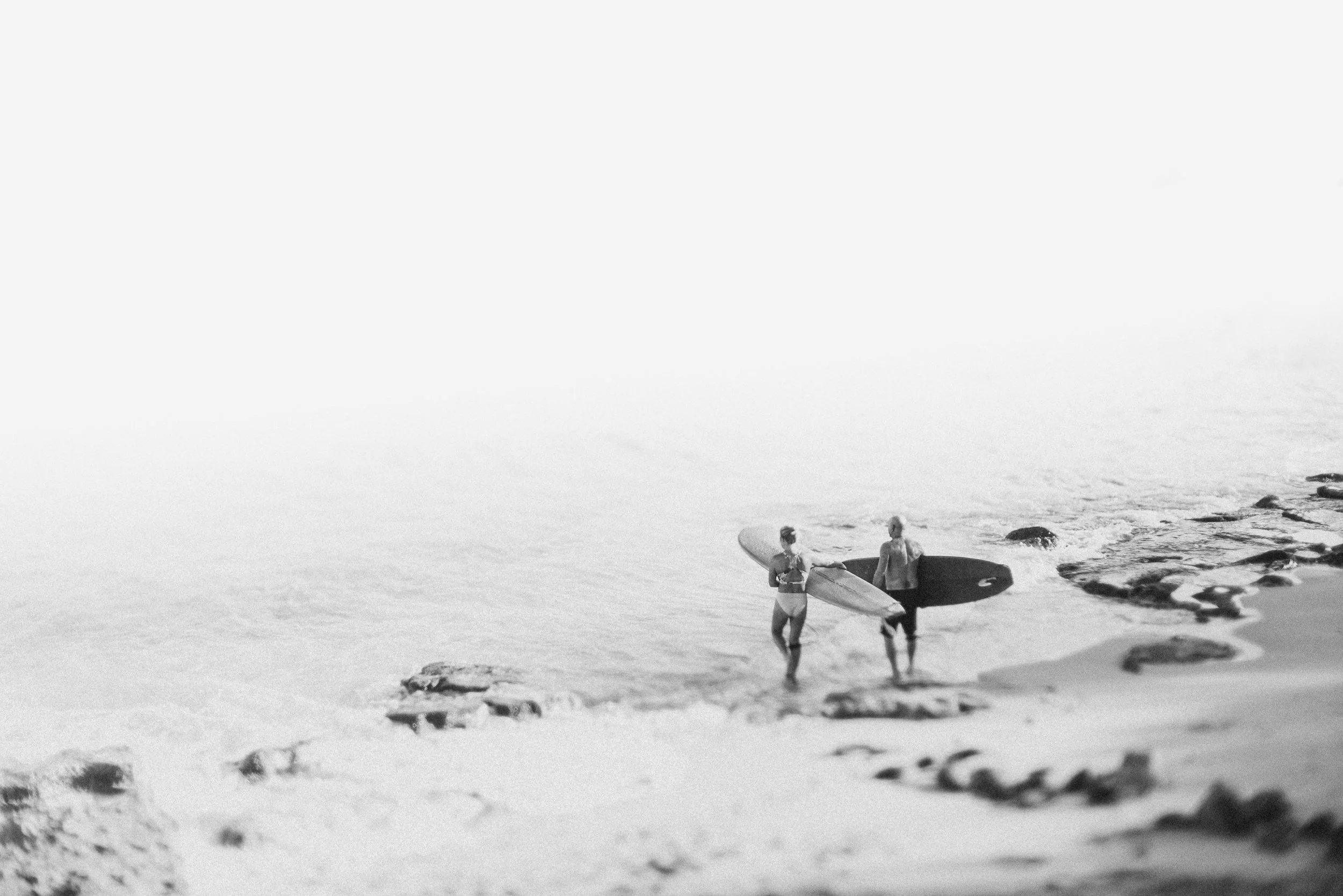 Two women with surfboards walking on the beach near rocks, with the ocean in the background.