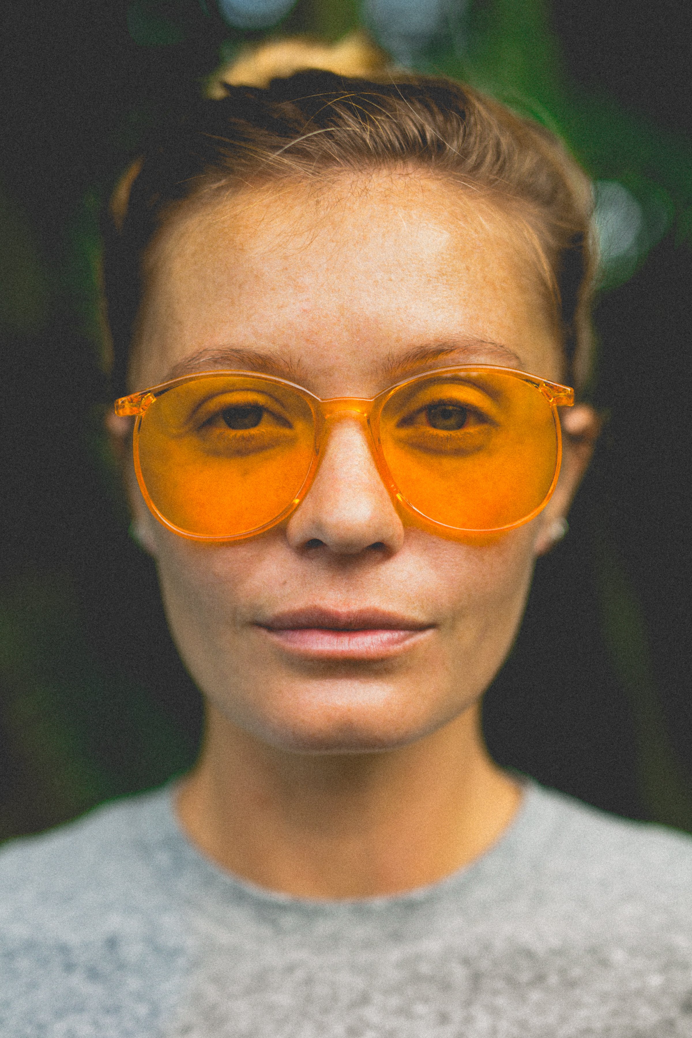Close-up of a woman wearing orange-tinted sunglasses, with a background of green foliage. She has short, light brown hair and a neutral facial expression.