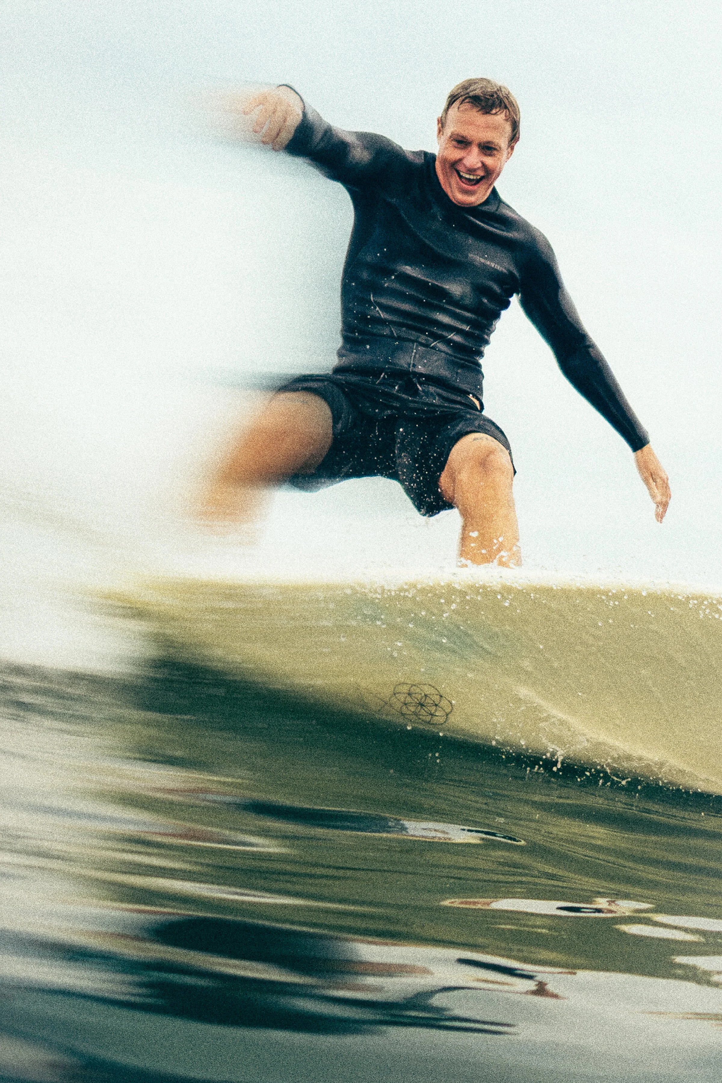 Man riding a surfboard on a wave, smiling, wearing a wetsuit.