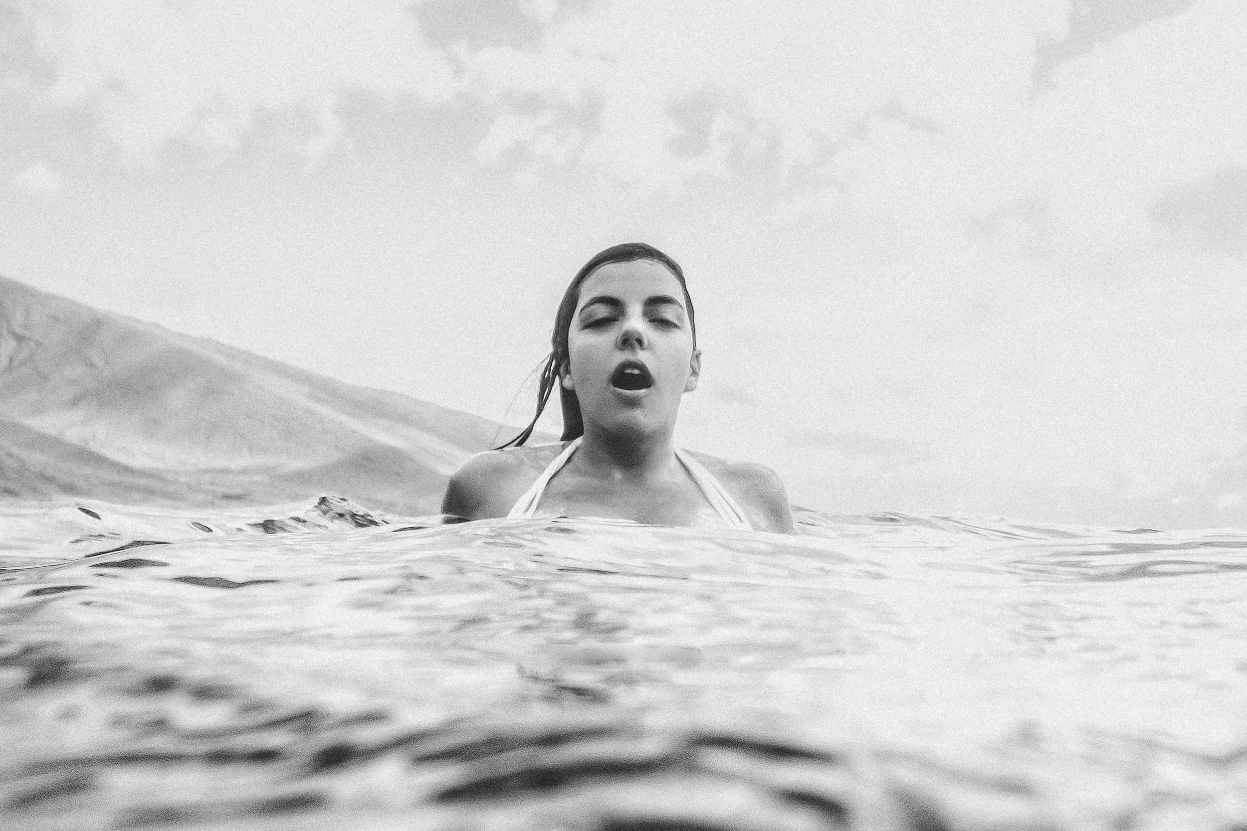 A woman with wet hair in a swimsuit emerging from the water with a mountain landscape in the background.