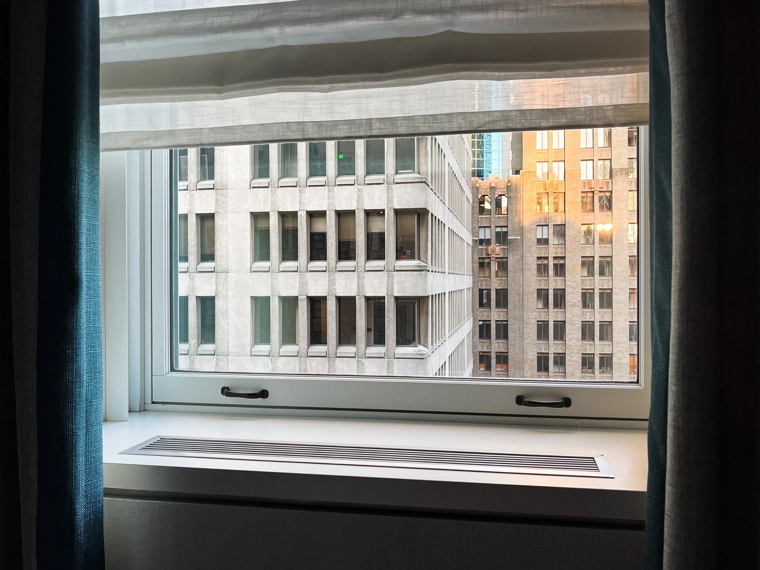 View of a city street seen through a window with blue curtains. The buildings are modern, with many windows and some reflective surfaces, and are bathed in warm sunlight.